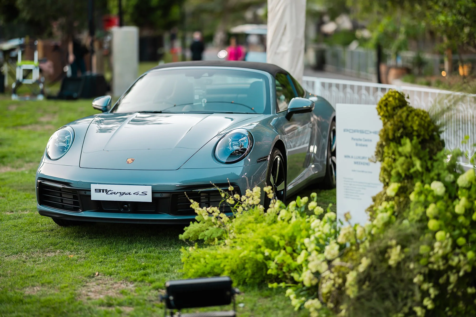 A silver Porsche 911 Targa 4S sports car displayed outdoors on a grassy area with a signboard and green flowering bushes in front.