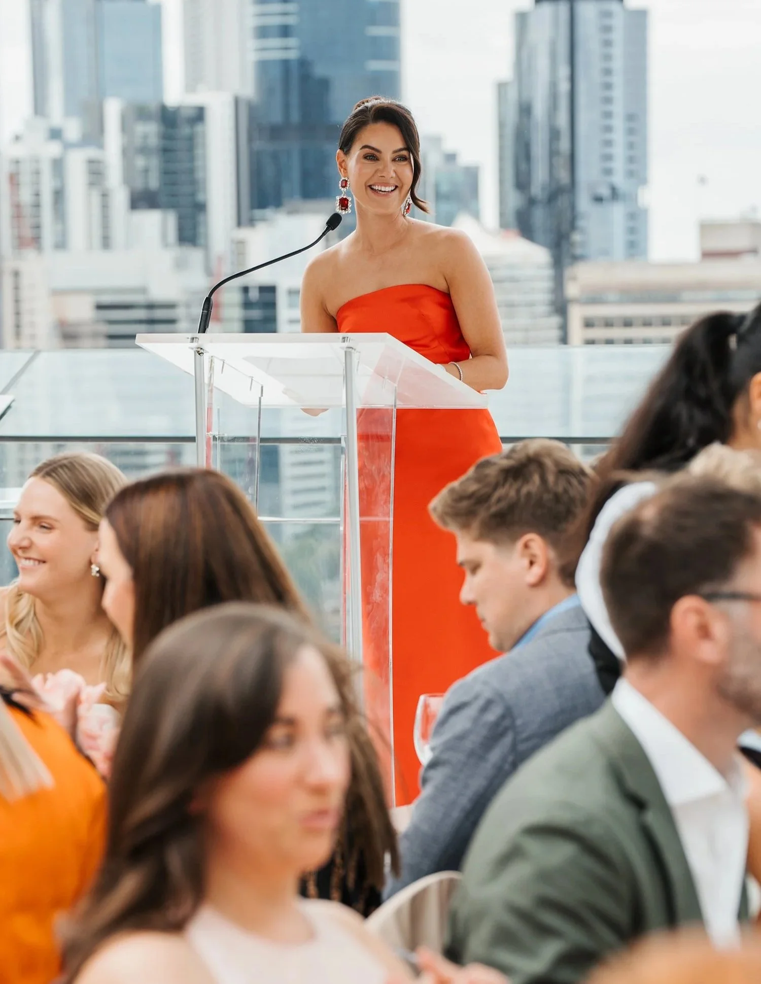 A woman in a red strapless dress speaking at a podium during a formal event in a city with tall buildings in the background.