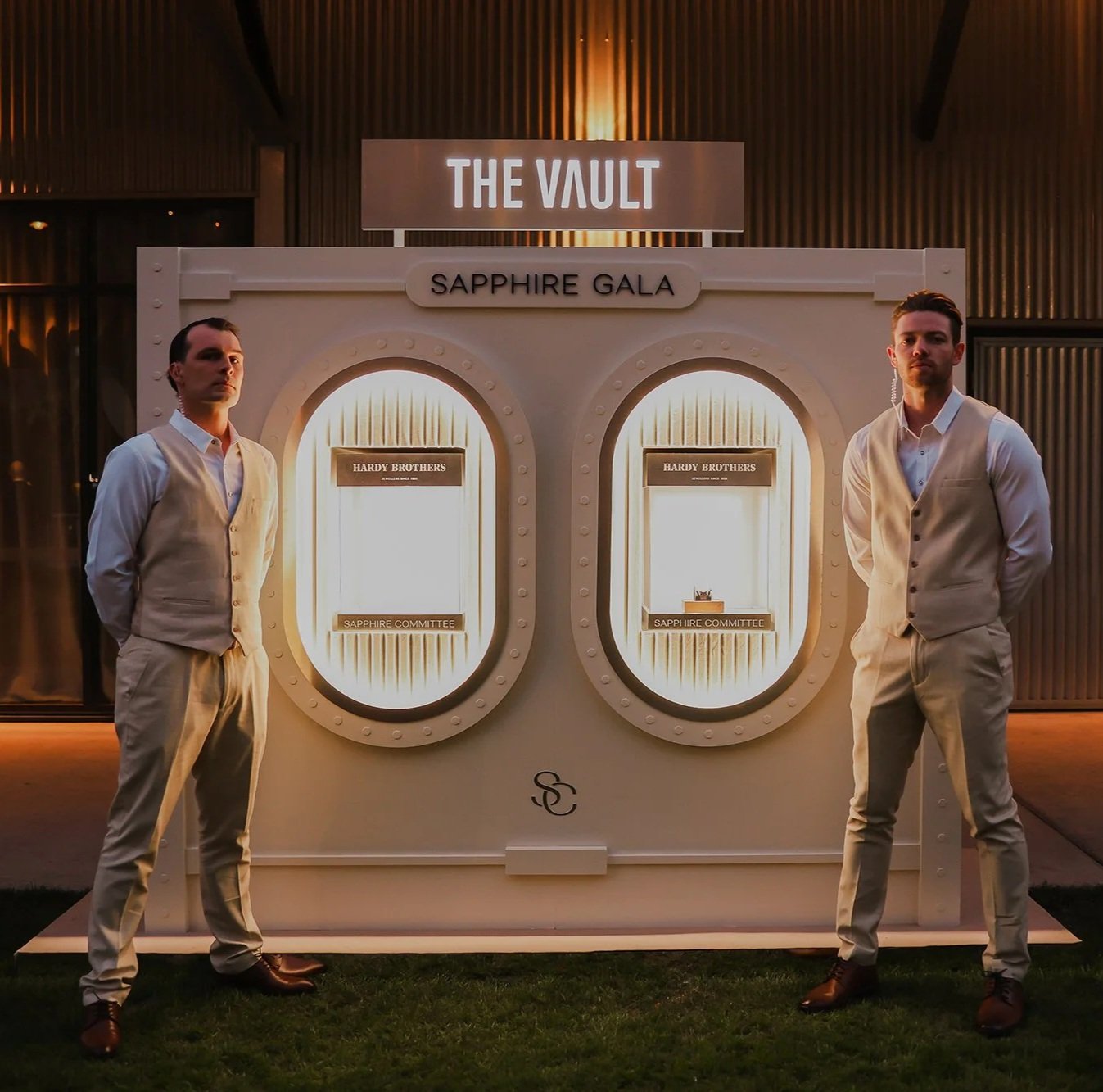 Two men in light-colored vests and trousers standing on either side of a white display booth with lit oval windows, labeled 'The Vault' and 'Sapphire Gala', with a sign for the 'Hardy Brothers' and 'Sapphire Committee,' in a dimly lit outdoor setting.