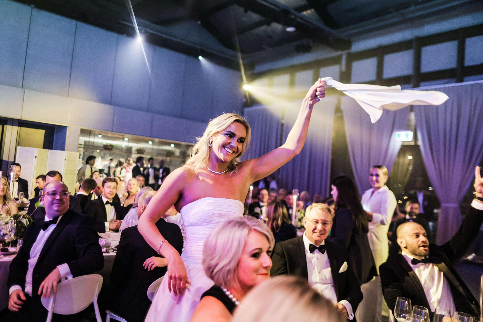 A woman in a white strapless dress is waving a white cloth in the air at a formal event, surrounded by seated guests in tuxedos and evening dresses.