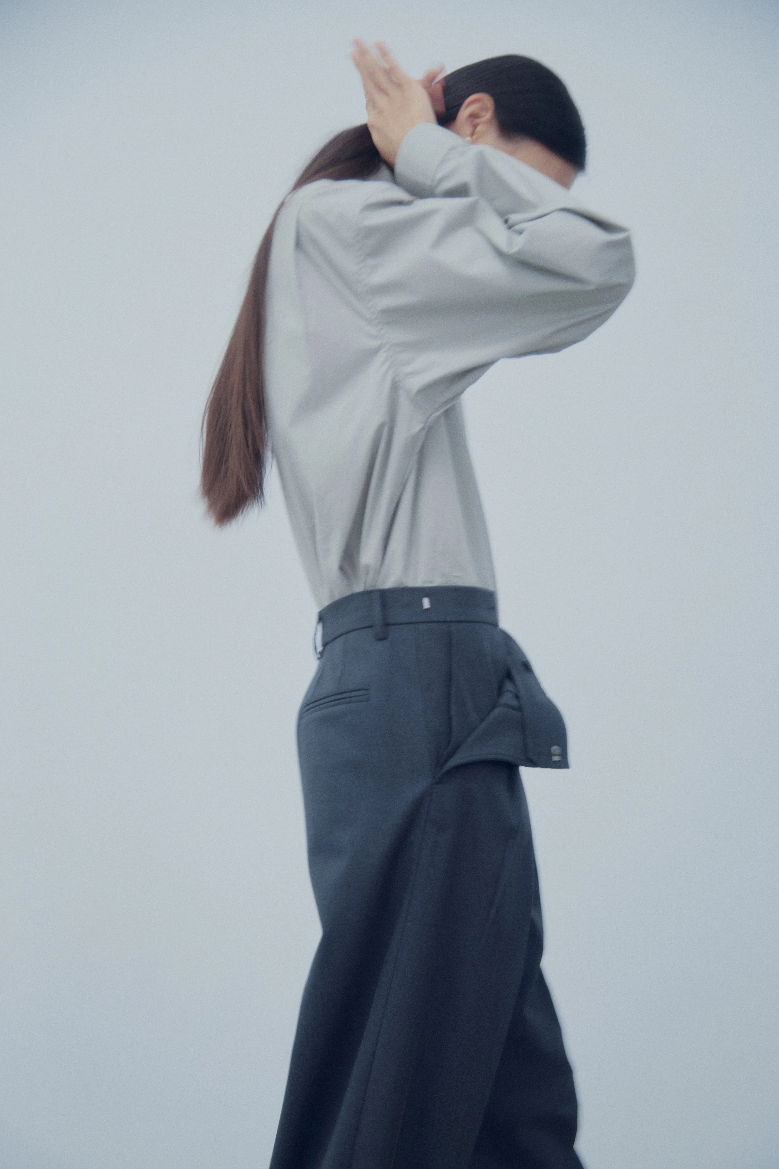 Person in gray shirt and dark pants with long hair covering face, standing against a plain background.