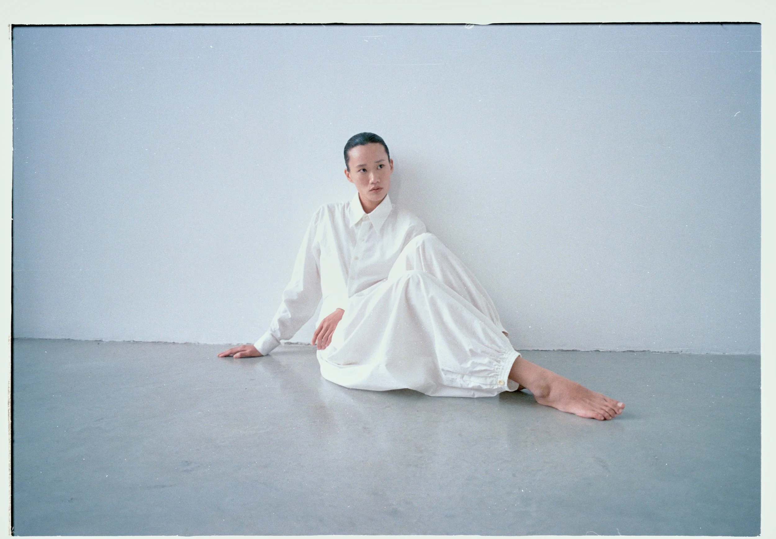 Person sitting on floor in white shirt and pants against a white wall, with bare feet.