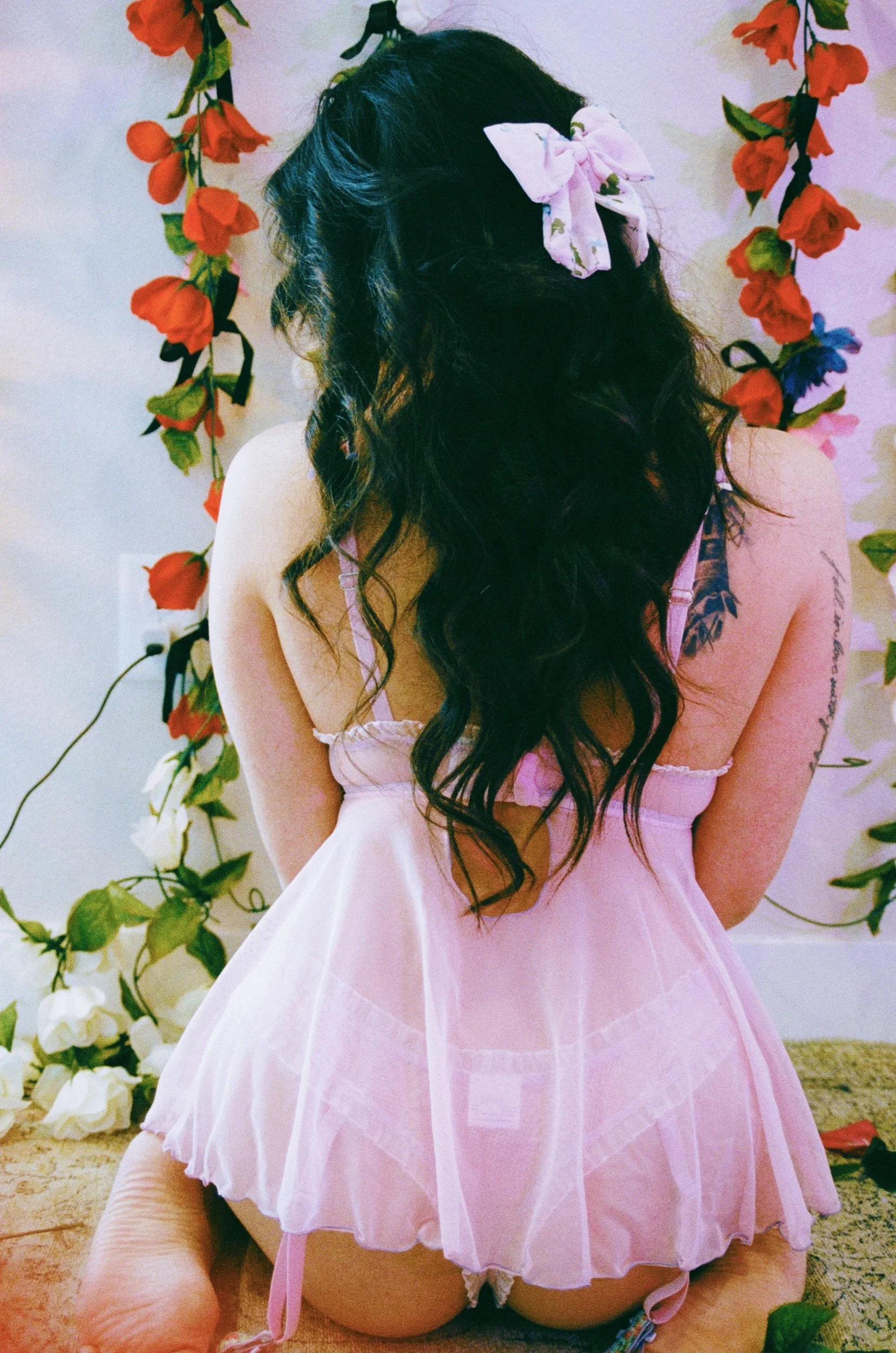 Woman with long hair and white bow in floral lingerie, rose-filled backdrop.