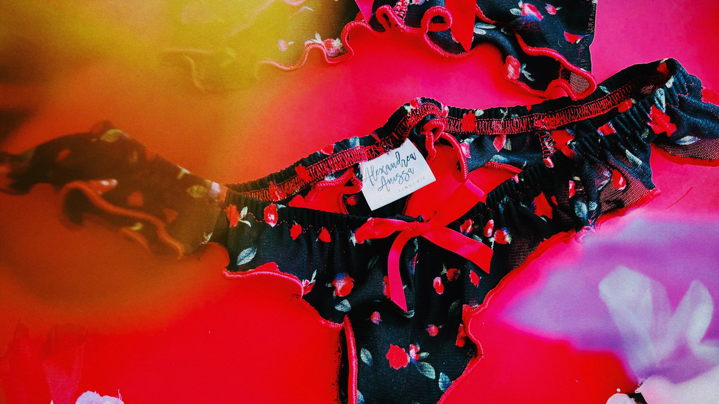 A close~up of the black floral lingerie set with red satin bow, styled against a vibrant floral arrangement and soft color contrasts.