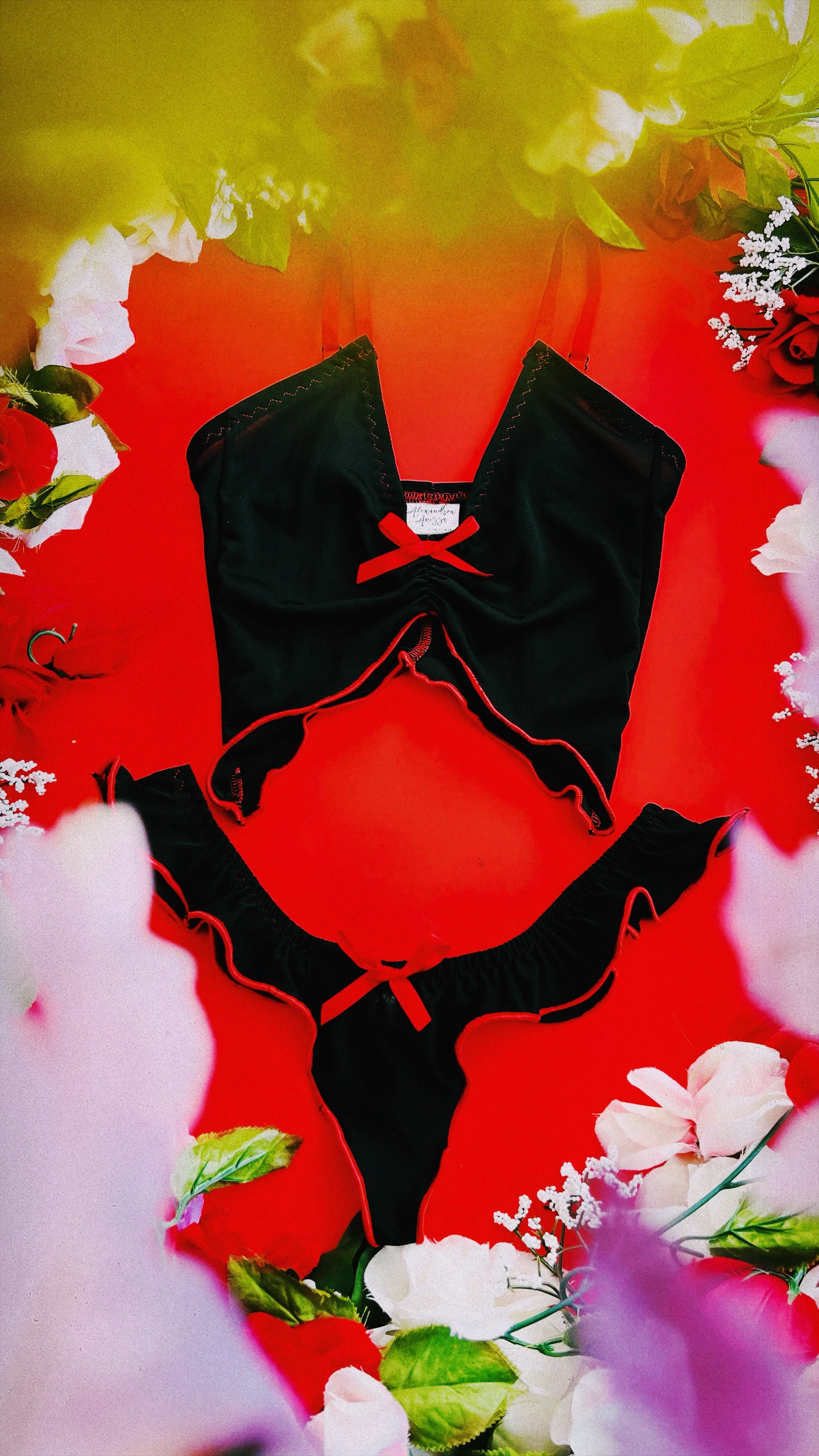 A two~piece black lingerie set with red rolled hems and bow accents, displayed against a vibrant red background surrounded by soft floral decor.