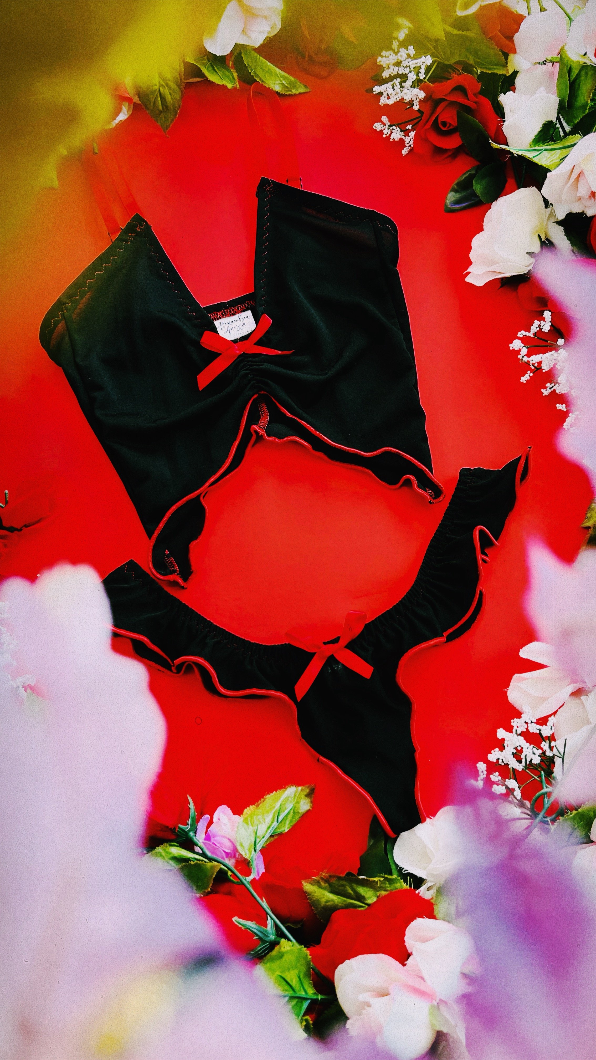 A two~piece black lingerie set with red rolled hems and bow accents, displayed against a vibrant red background surrounded by soft floral decor.