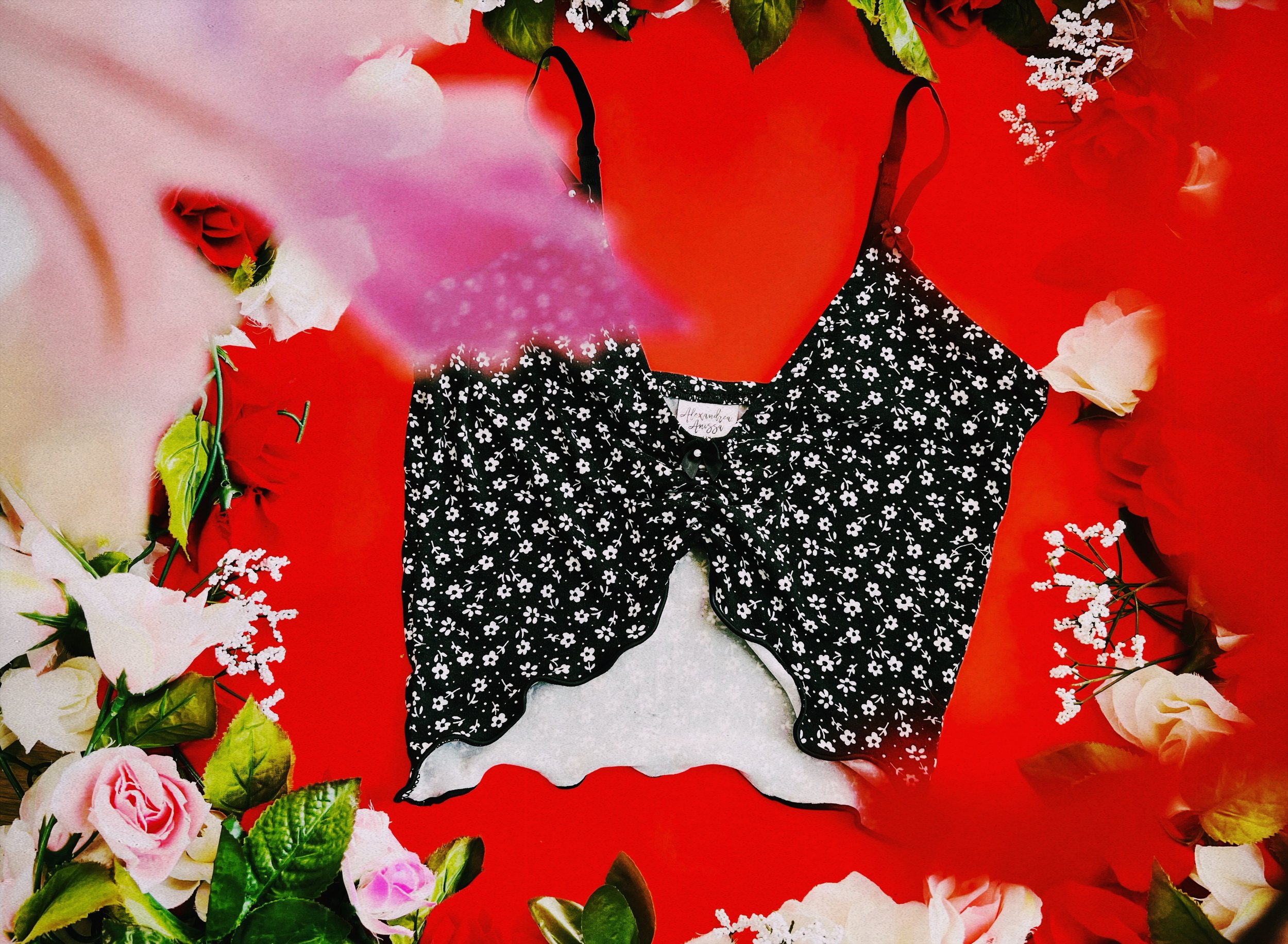 A lightweight black cami top with a white floral print and scalloped hemline, displayed against a vibrant red backdrop adorned with roses and greenery.