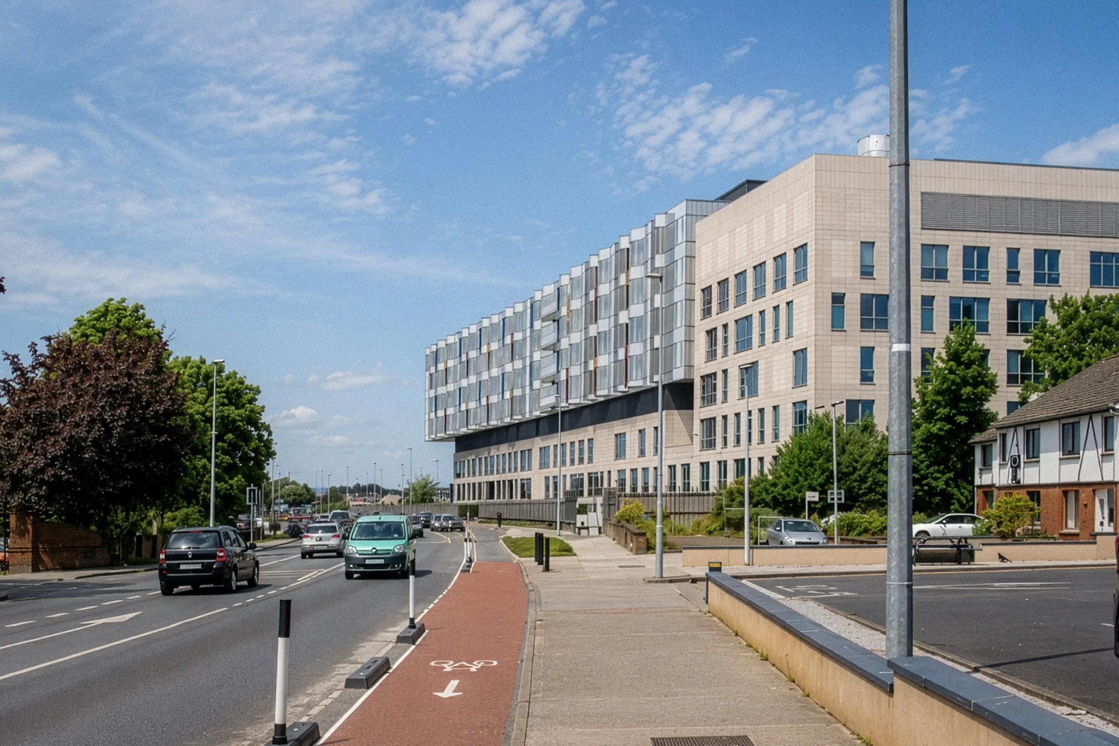 Urban street scene with parking lot, modern multi-story building, trees, and a blue sky with scattered clouds.