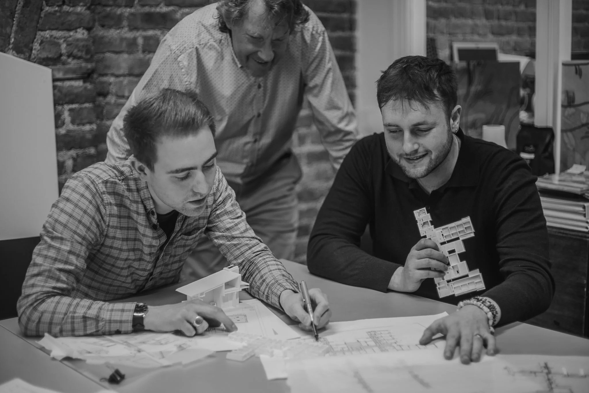 A group of people working together on architectural plans and models at a table in a room with a brick wall. One person is showing models to others, and they appear engaged in discussion.