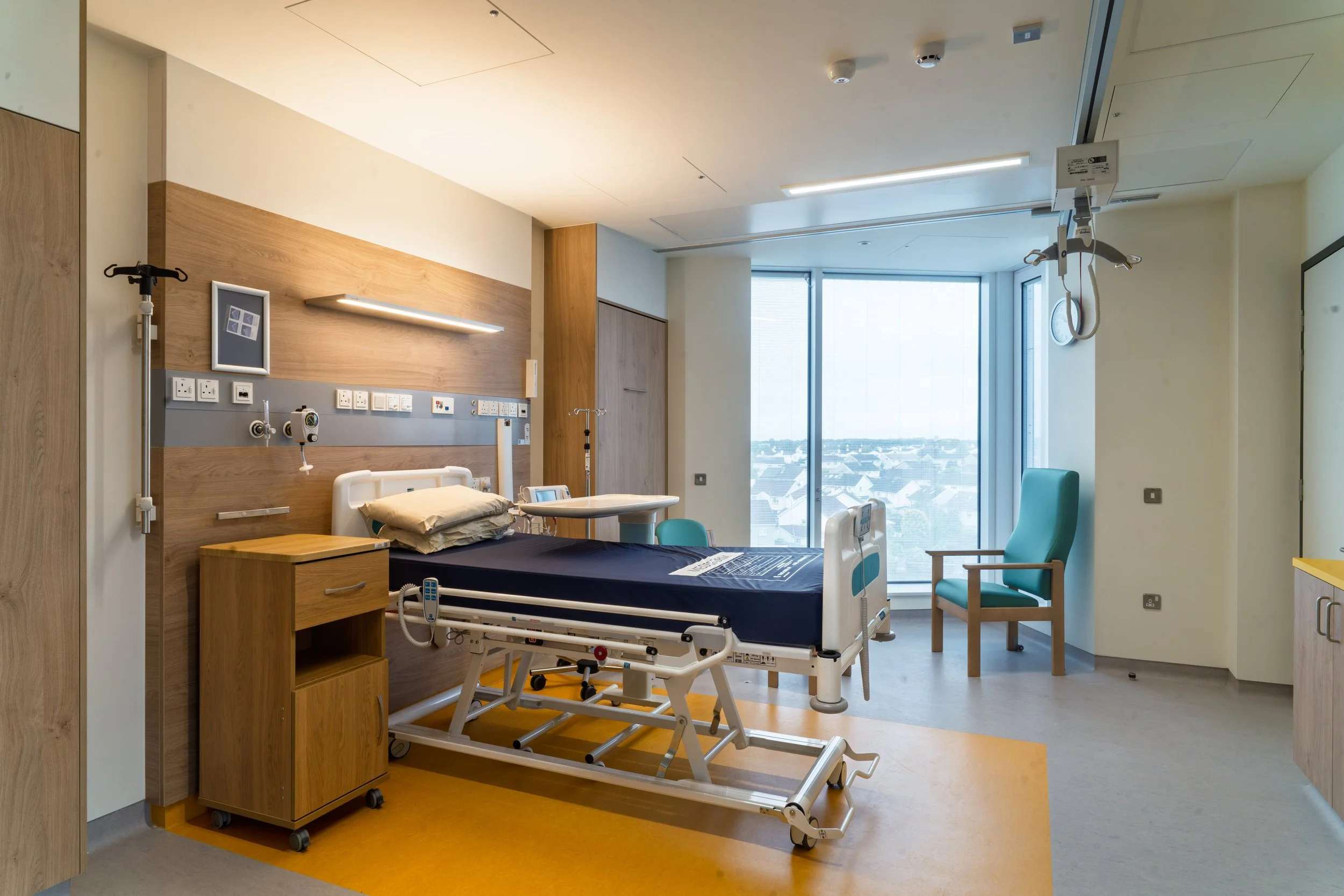 Hospital room with a hospital bed, medical equipment, a wooden nightstand, a blue chair, and large windows overlooking a cityscape.