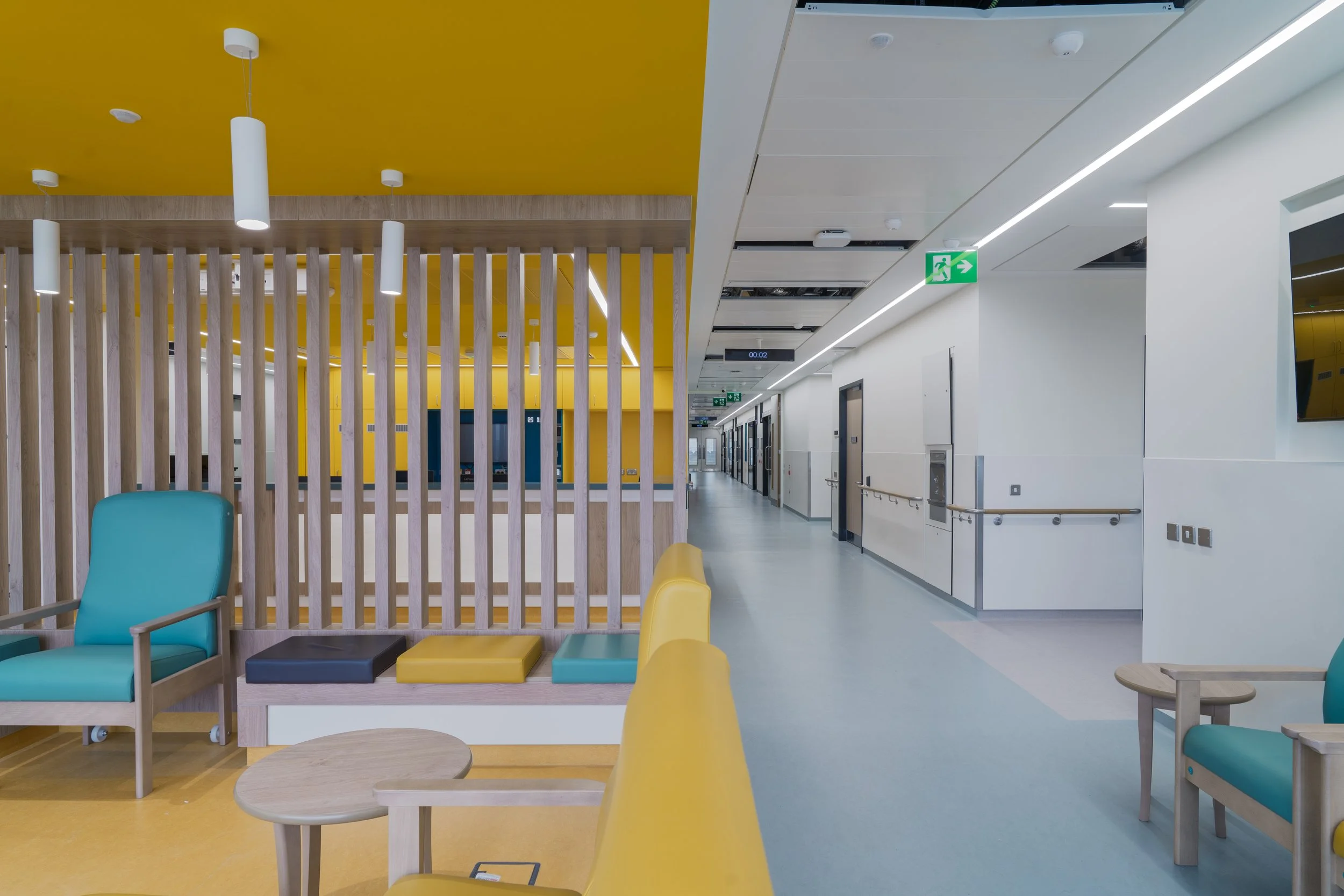 Interior of a modern building with yellow and white walls, wooden and colorful furniture, and ceiling lights. Emergency exit sign is visible.