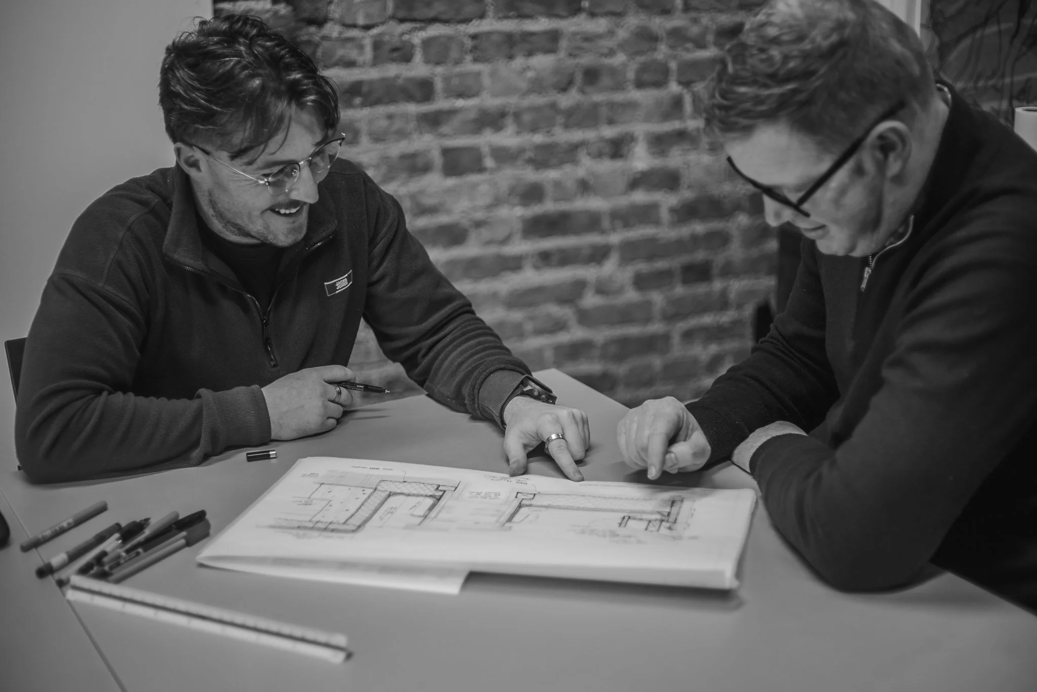 Two men are sitting at a table, closely examining and discussing architectural blueprints. One man is pointing to a specific area on the blueprint as they both look at it attentively. The table is scattered with markers and additional papers. The background features an exposed brick wall.
