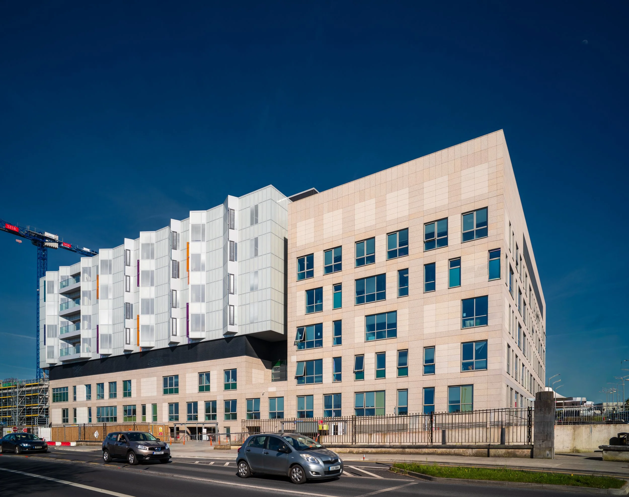 Modern multi-story building with a unique architectural design, large windows, and a crane nearby under a clear blue sky, with cars parked in the foreground.