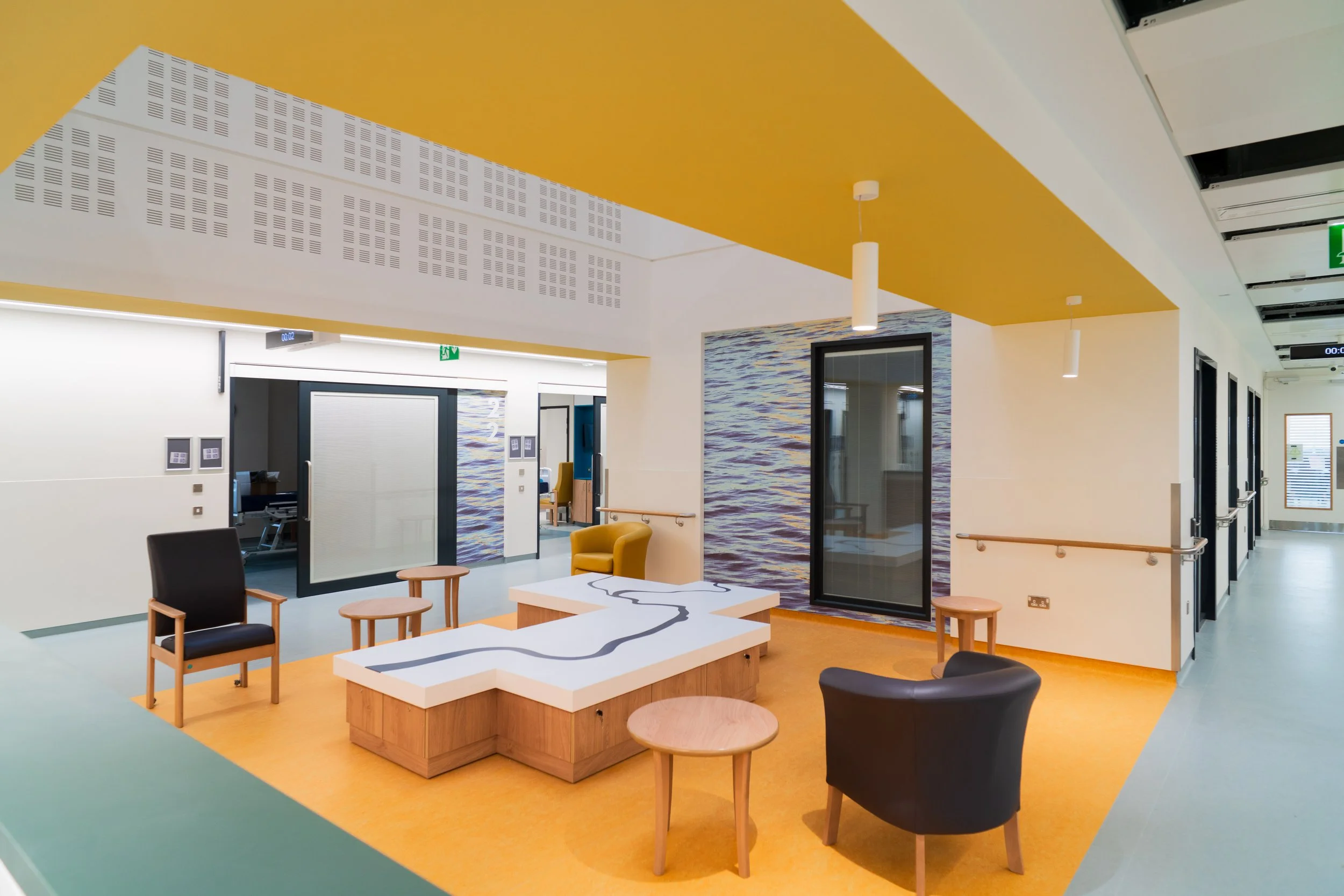 Modern interior space with yellow and green flooring, white walls, black chairs, wooden tables, and ceiling lights, featuring large artworks and glass doors.