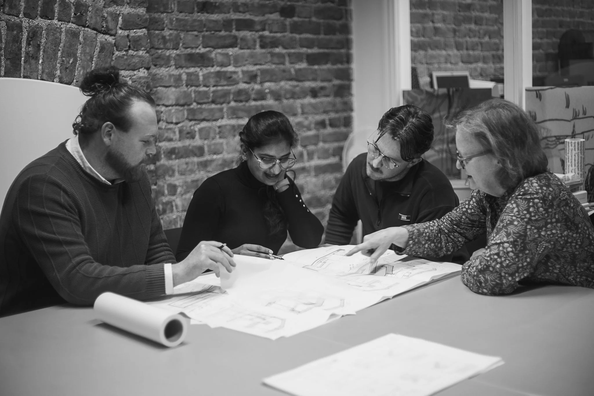 Group of four people sitting at a table, engaged in discussion over blueprints or architectural plans in an office with brick walls.