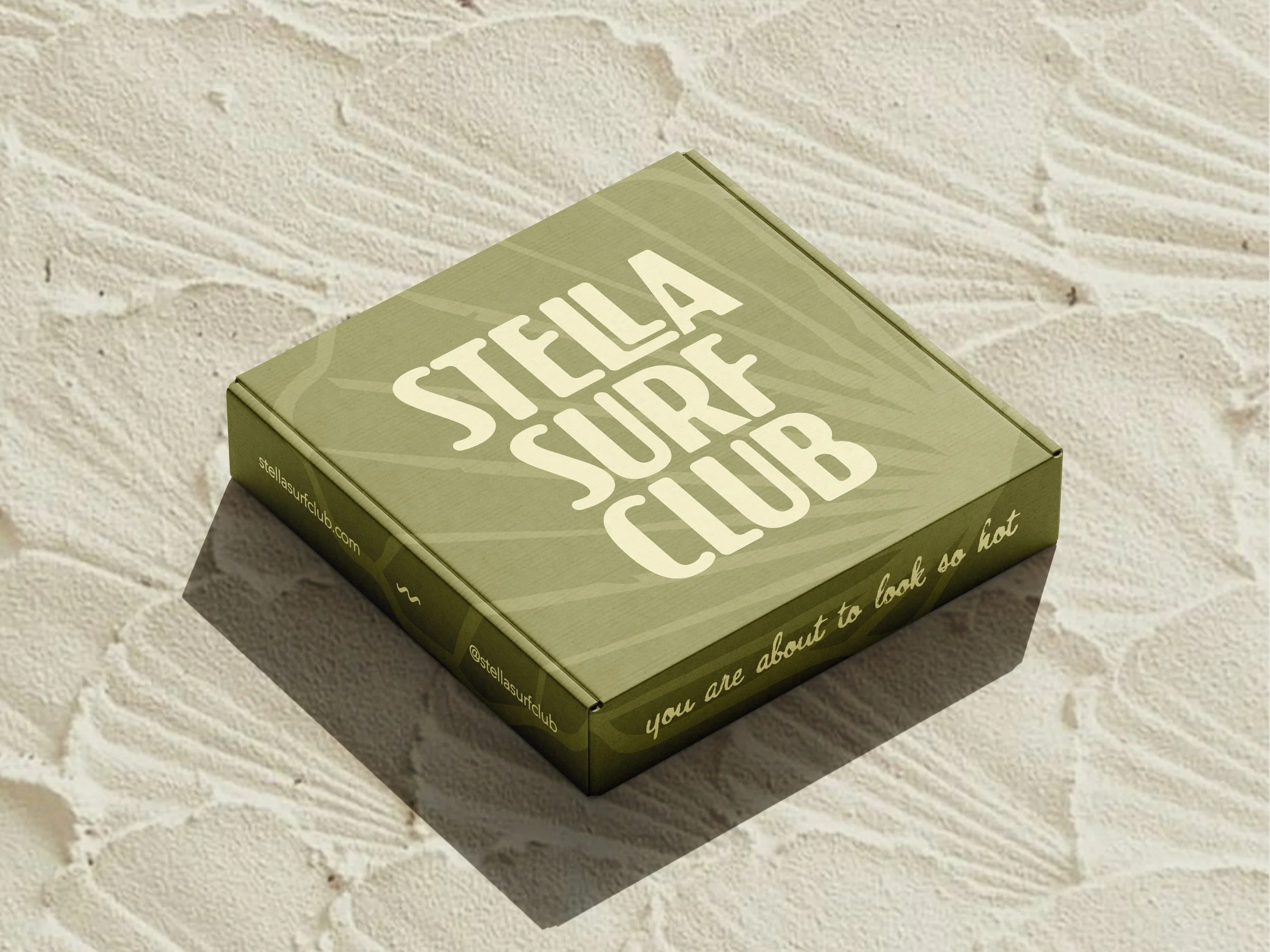 A green box titled 'Stella's Surf Club' is placed against a light-colored textured wall. The box has some website or social media handle printed on the side.