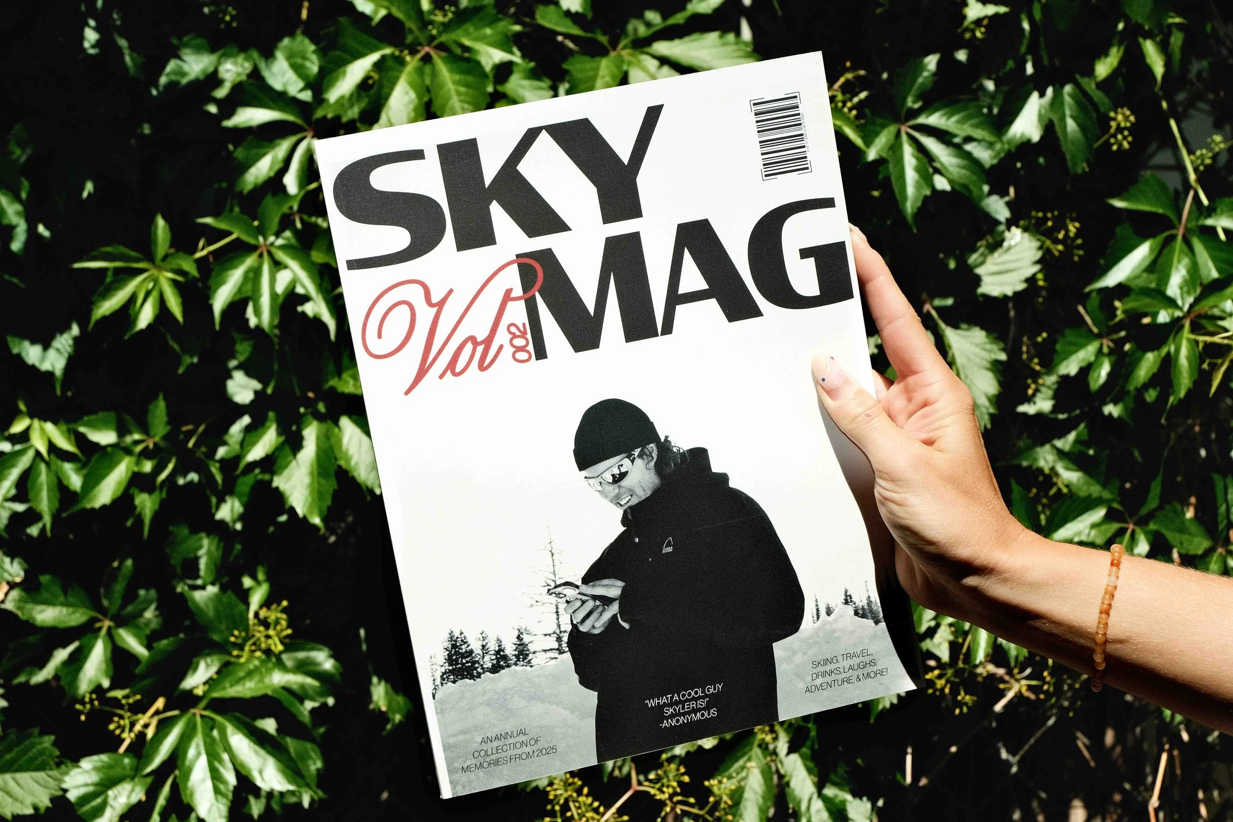 A person holding a magazine with a black and white cover image of a man in winter clothing, with a background of green leafy plants.