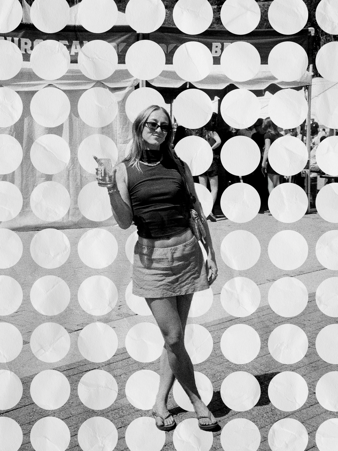 A woman posing at an outdoor event, holding a drink, wearing sunglasses, a sleeveless top, a skirt, and sandals, with tents and people in the background.