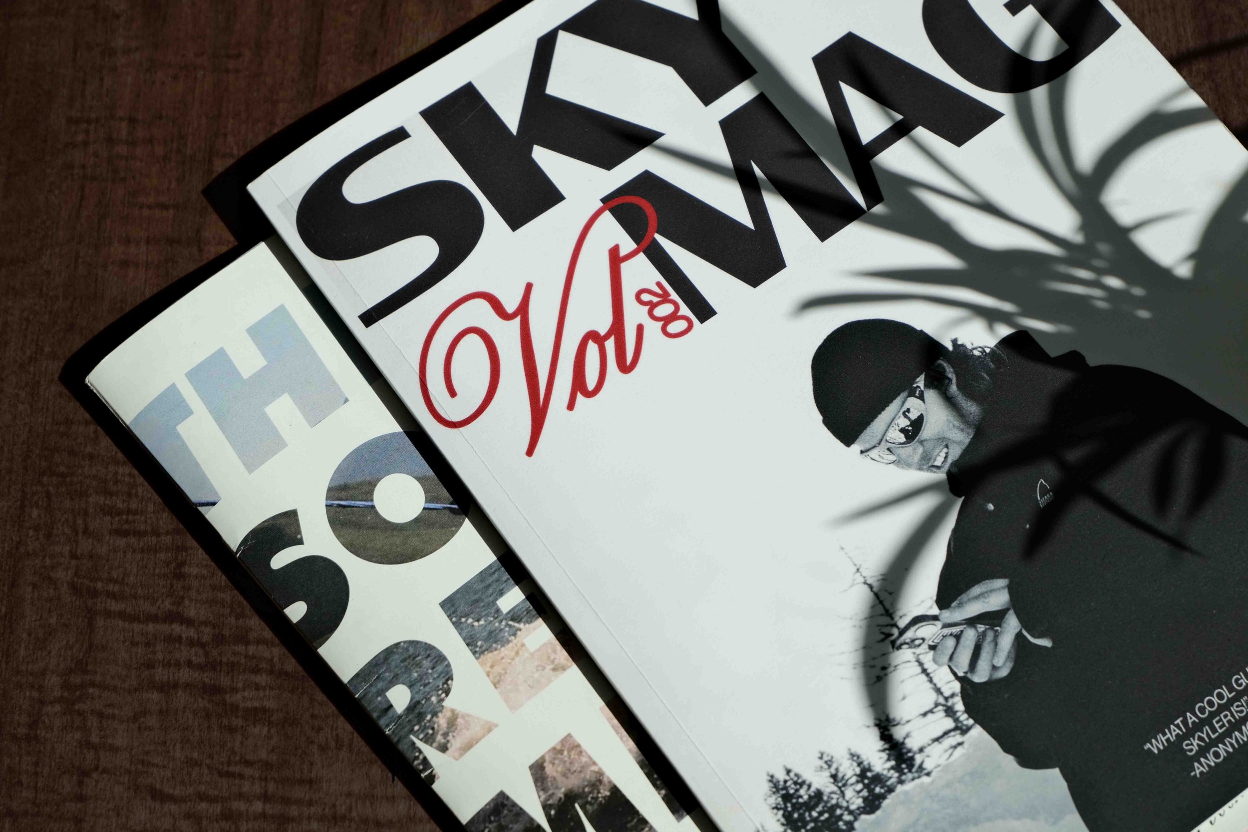 A collection of magazines stacked on a dark wooden surface, with a shadow of a plant cast over the top magazine. The magazines feature bold text and images on their covers.