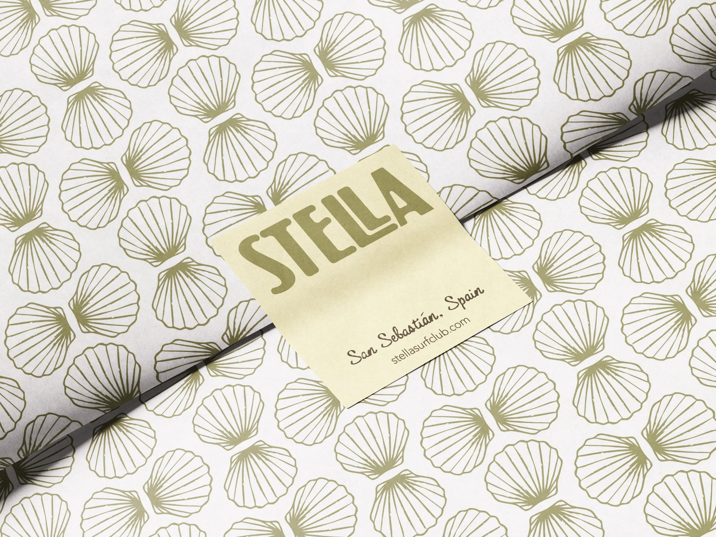 Close-up of patterned wrapping paper with gold seashell designs, partially covered by a yellow note labeled 'SELLA' and the text 'San Sebastian, Spain' and a website address.