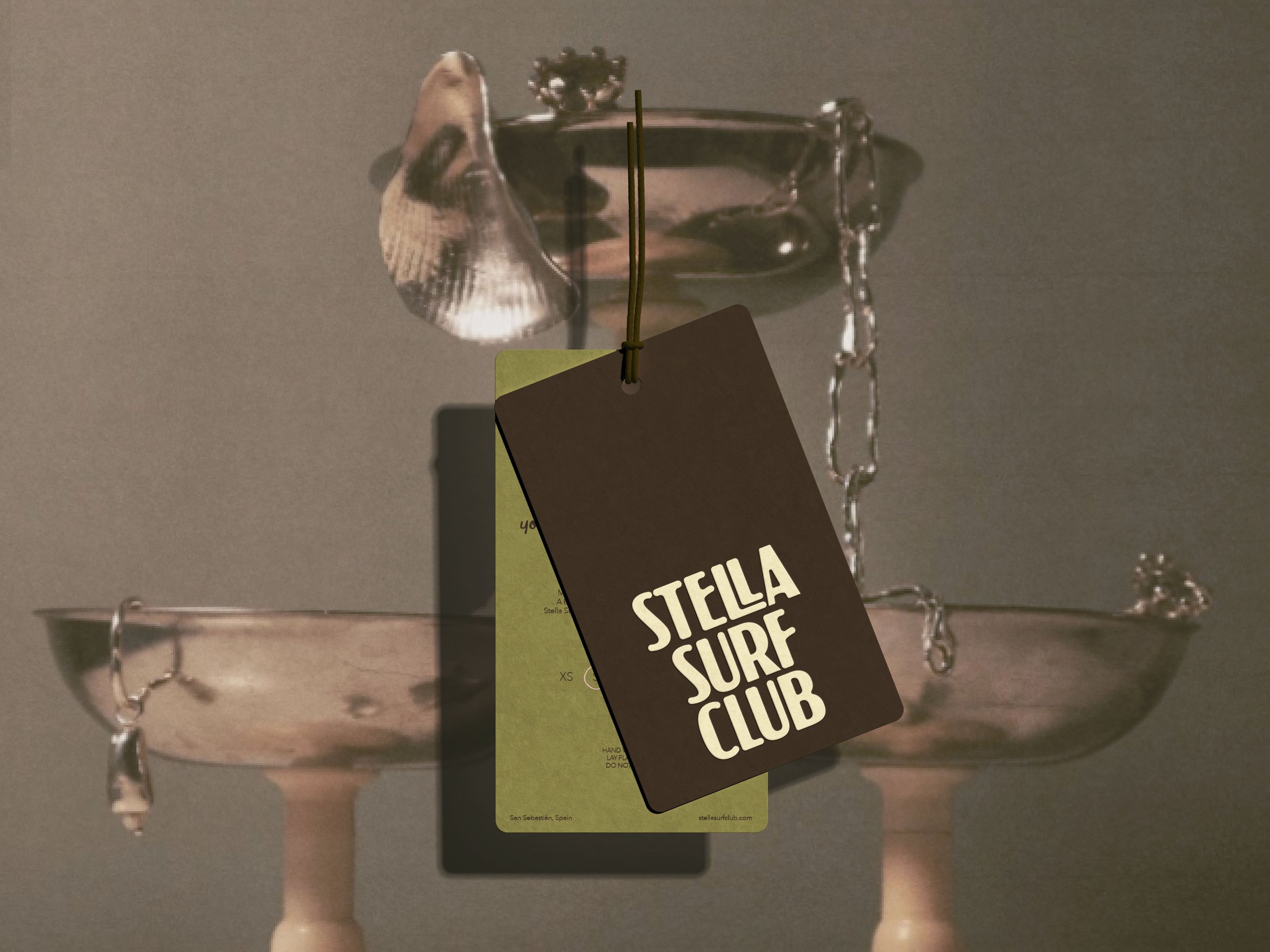 A metallic bowl with a chain attached and two tags hanging from it, one black labeled "Stella Surf Club" and one green tag. The scene appears to be indoors with a gray background.