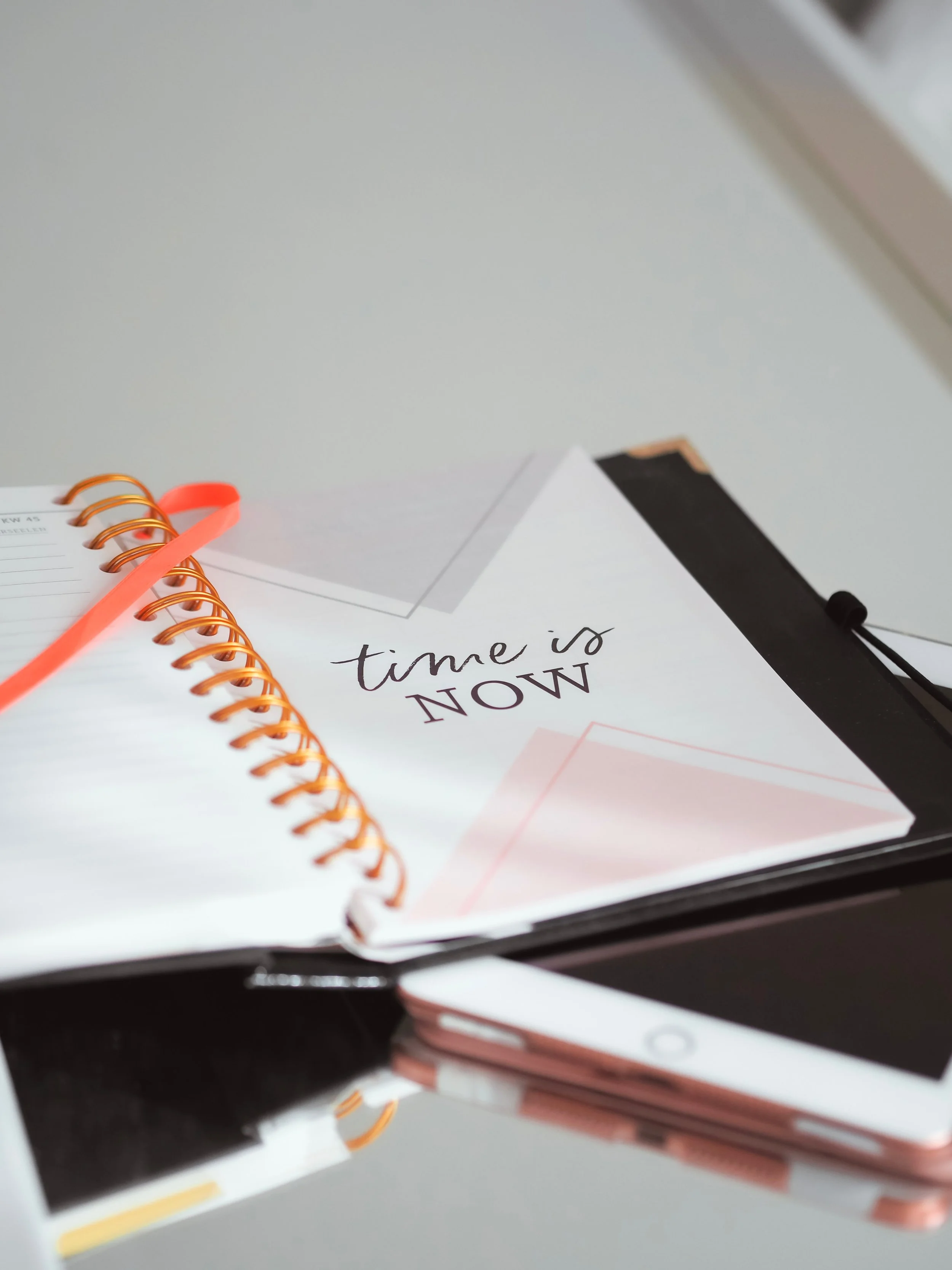 Open notebook with "time is NOW" on the page, orange spiral binding, and an orange ribbon bookmark, placed on a desk with a smartphone.