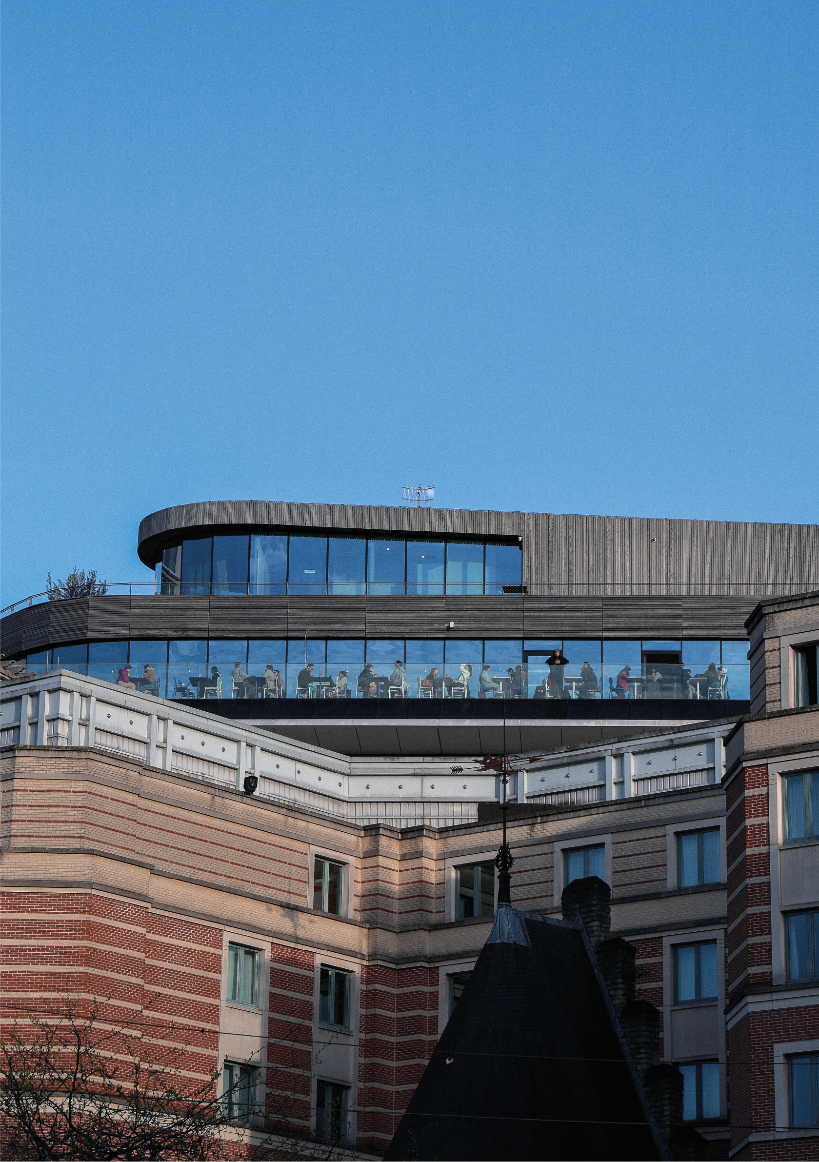 Vue d'un bâtiment moderne avec une terrasse en verre sur le toit, des personnes assises à des tables, sous un ciel bleu clair.