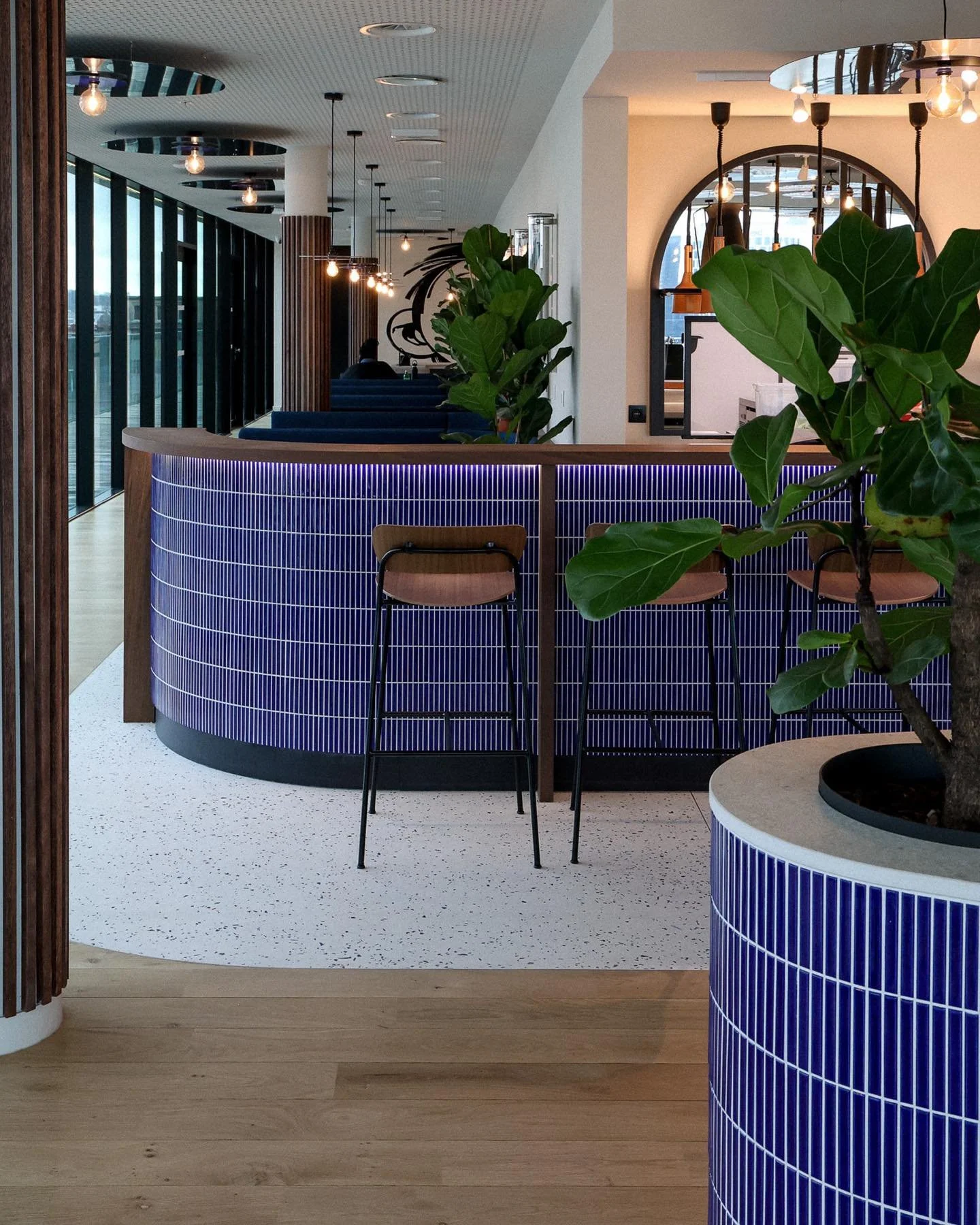 Intérieur d'un café ou restaurant moderne avec un comptoir en tuiles bleues, quelques chaises en bois, des plantes vertes et un éclairage chaleureux.