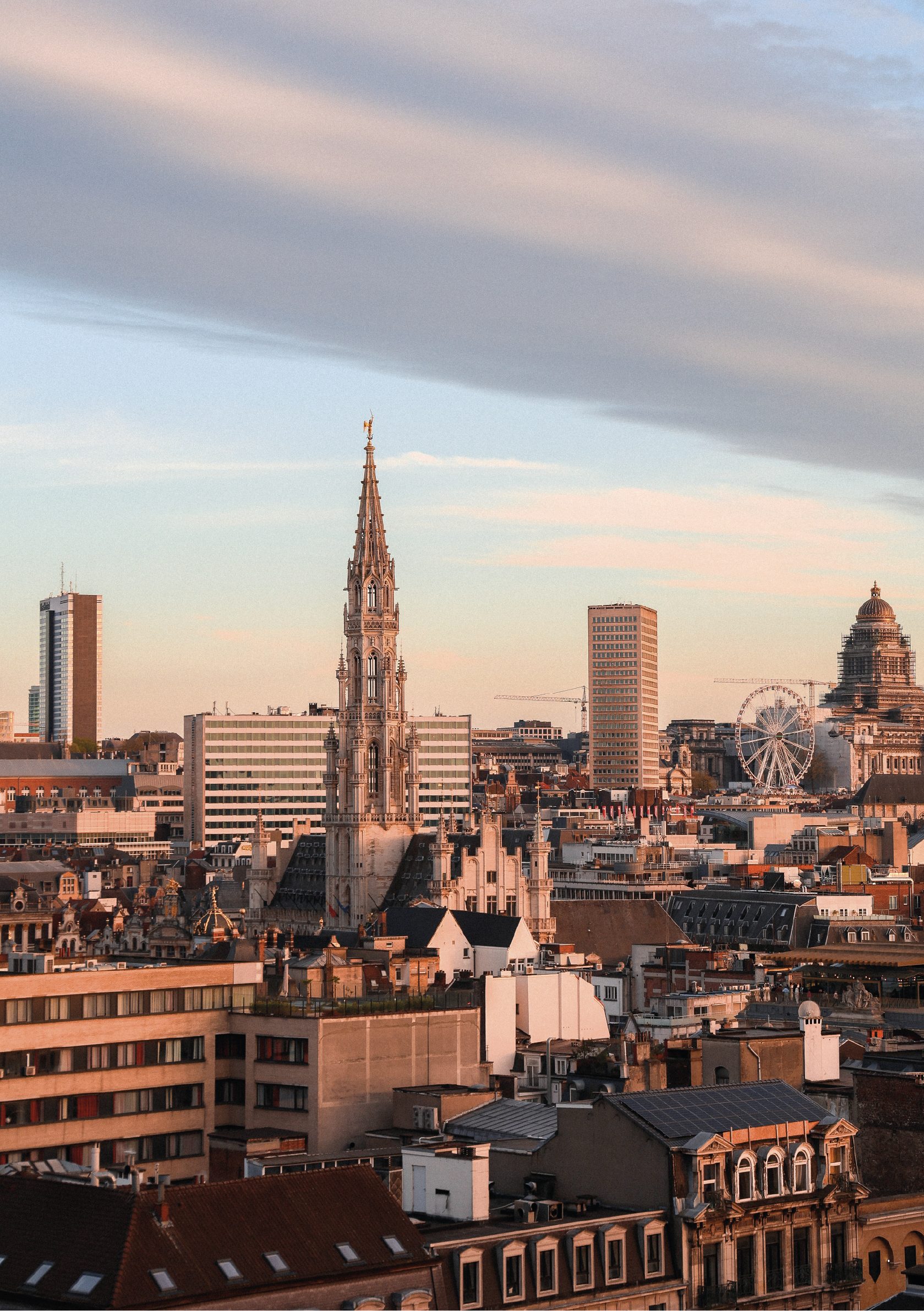 Paysage urbain avec des bâtiments anciens et modernes, une grande roue, et un ciel nuageux au coucher du soleil.