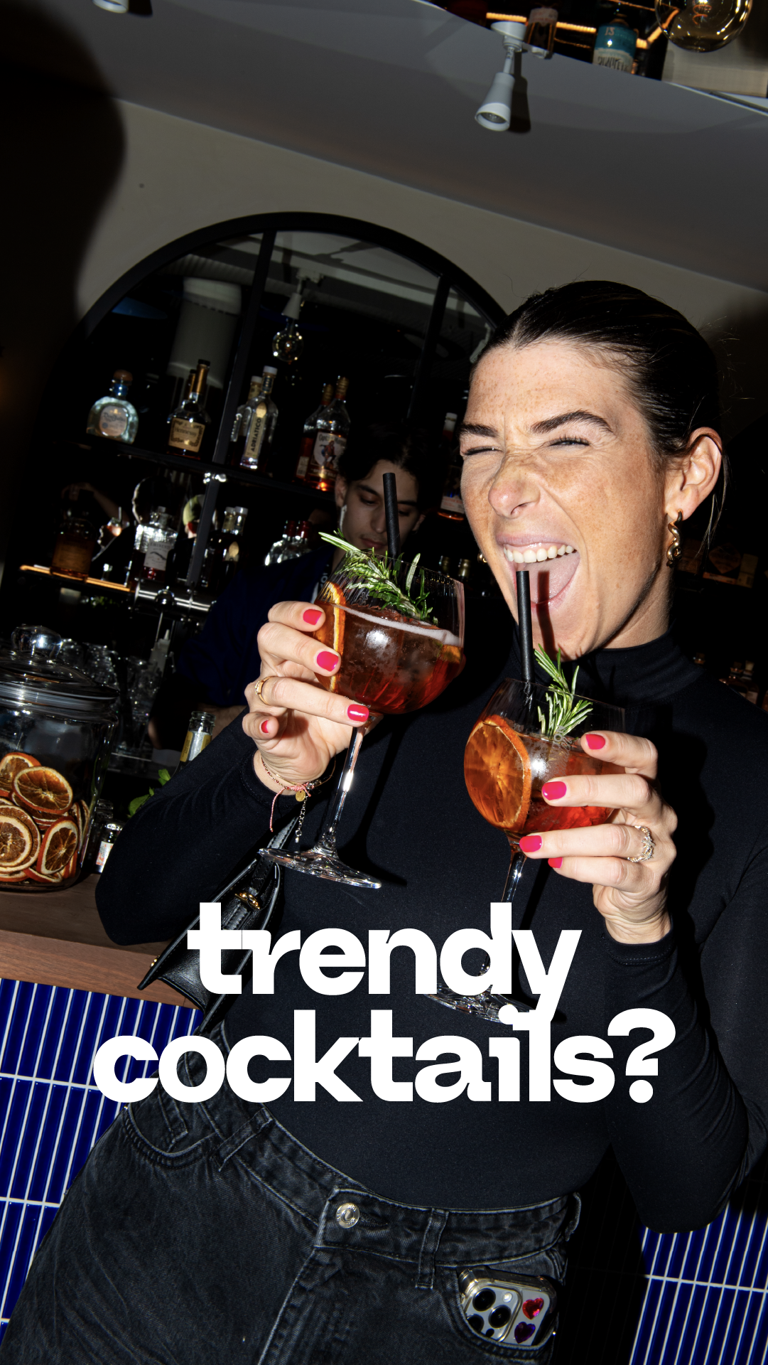 Une femme souriante en train de boire un cocktail dans un bar, portant un vêtement noir et des boucles d'oreilles, avec une étagère de bouteilles derrière elle.