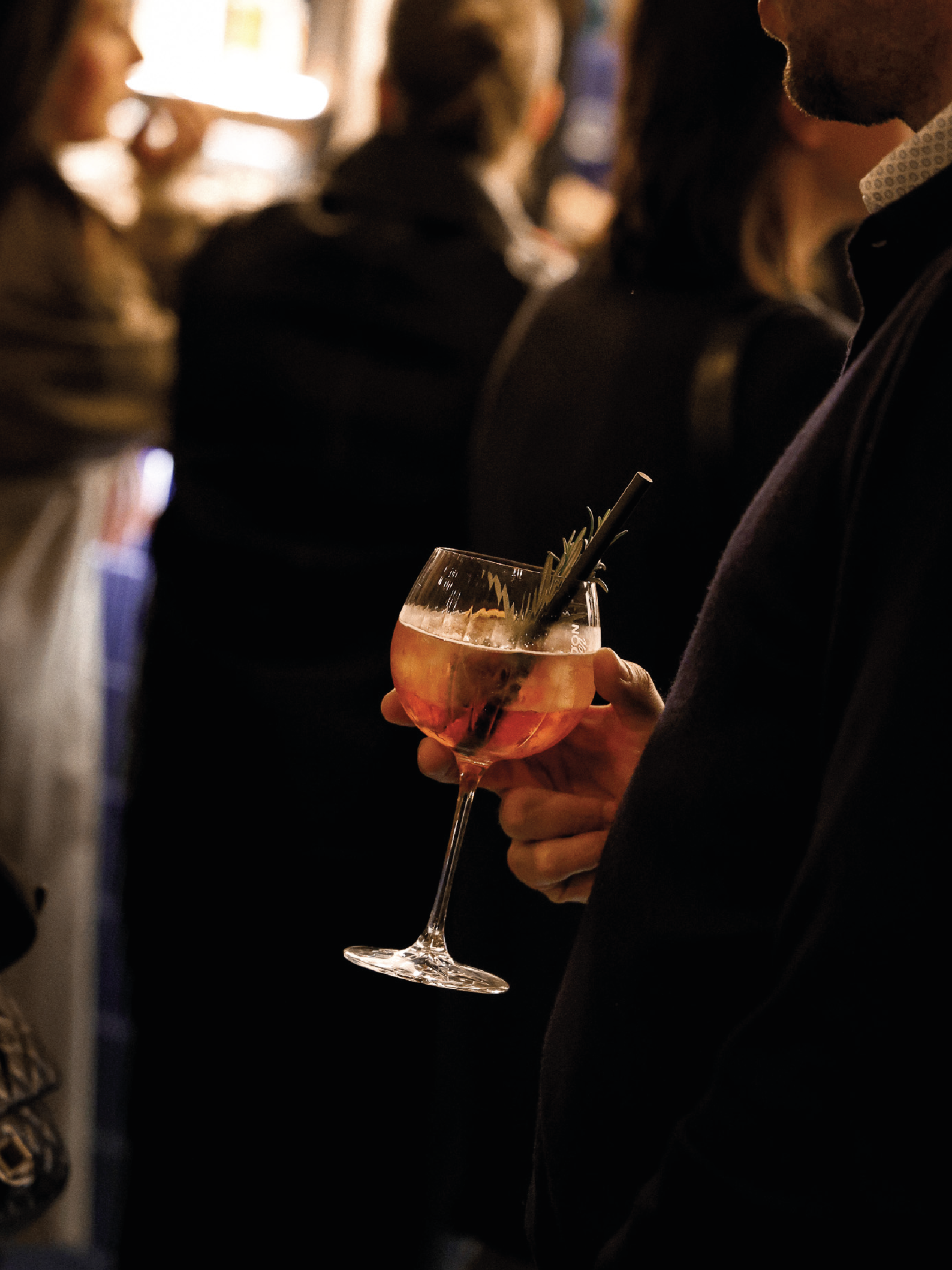 Une personne tenant un verre à cocktail translucide avec une boisson rose-orange, décorée d'une branche de romarin et d'une paille noire, dans un environnement de bar ou de fête avec d'autres personnes en arrière-plan.