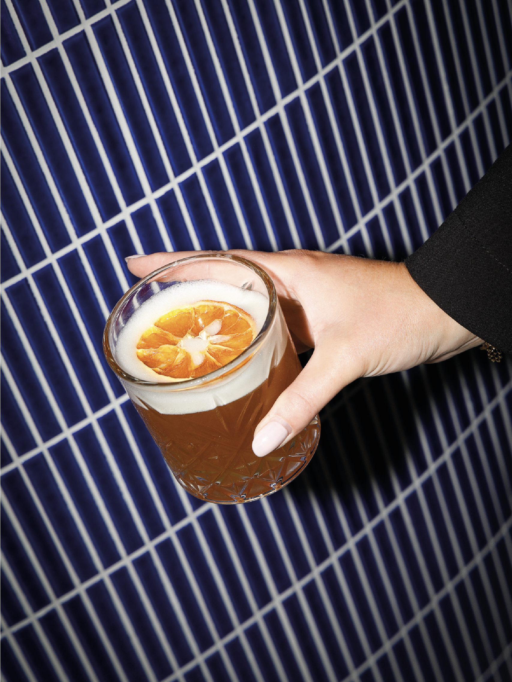 Un verre de cocktail avec une tranche d'orange sur le dessus, tenu par une personne dont la main porte une veste noire, sur un fond de carrelage bleu et blanc.