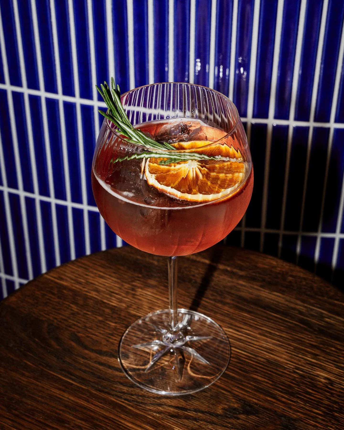 Verre à cocktail rose avec morceau d'orange, branche de romarin et glaçons sur une table en bois devant un mur en tuiles bleues