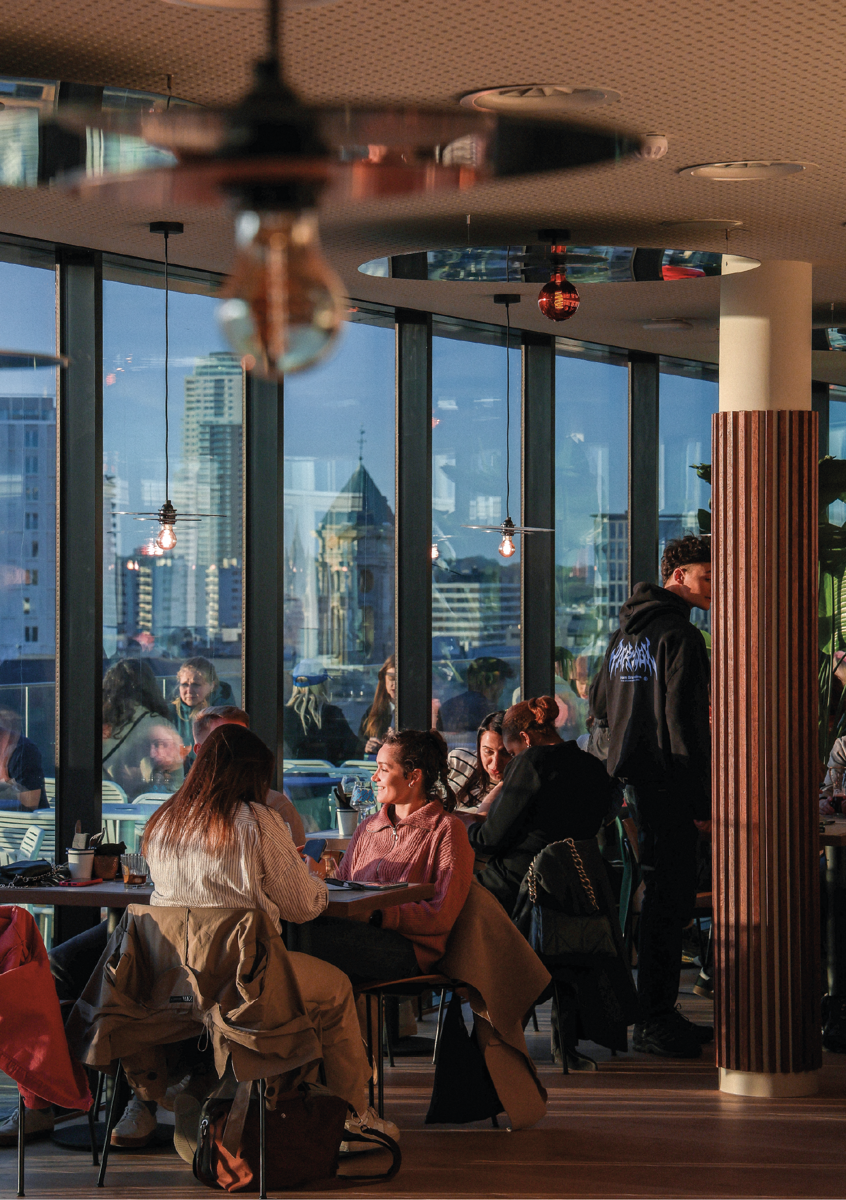 Groupe de personnes assises autour de tables dans un restaurant ou un café, avec de grandes fenêtres offrant une vue sur des bâtiments urbains. La lumière naturelle illumine la scène, créant une ambiance conviviale.