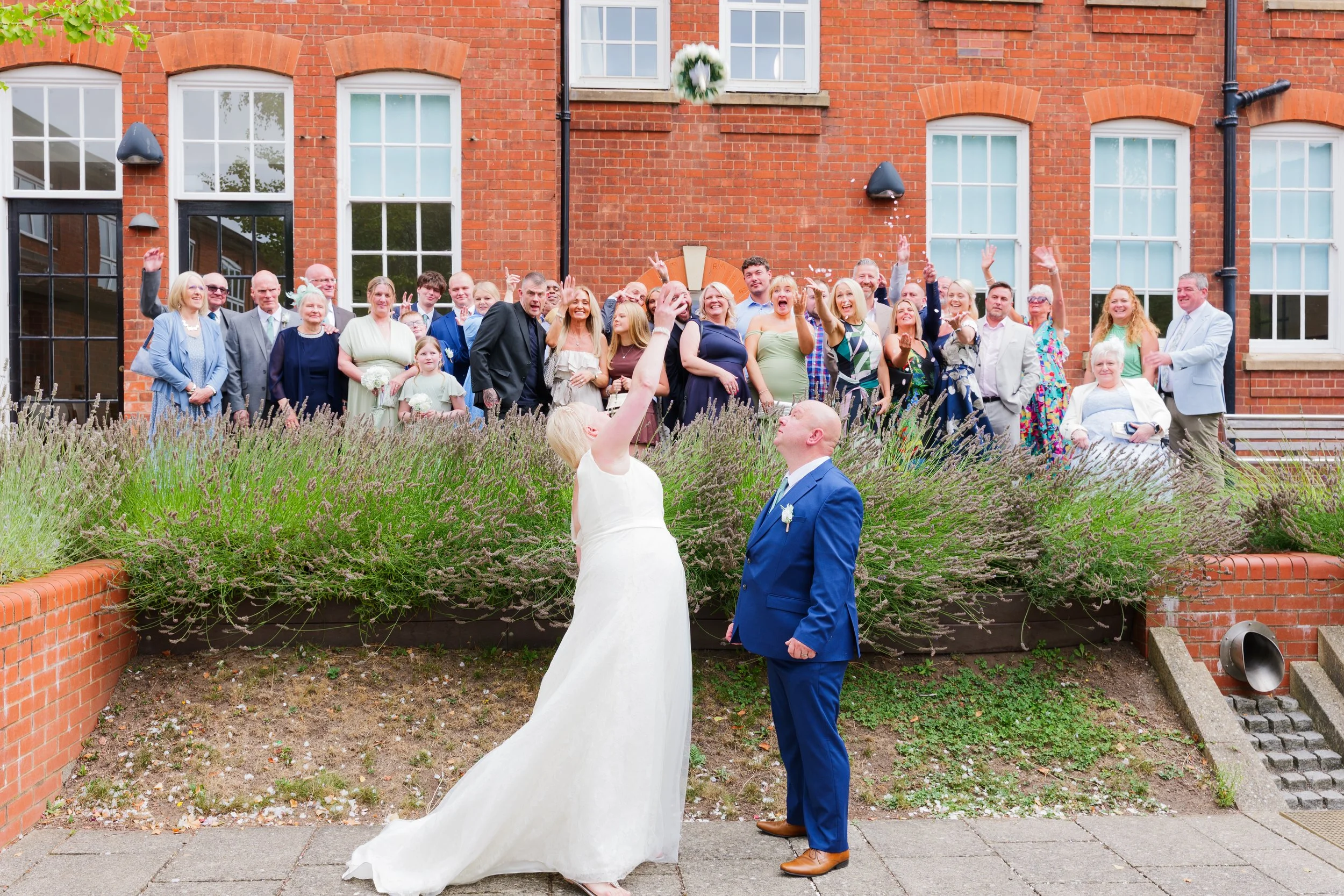 A bride and groom stand outside in front of a brick building during their wedding ceremony, with a large group of guests behind them, celebrating.