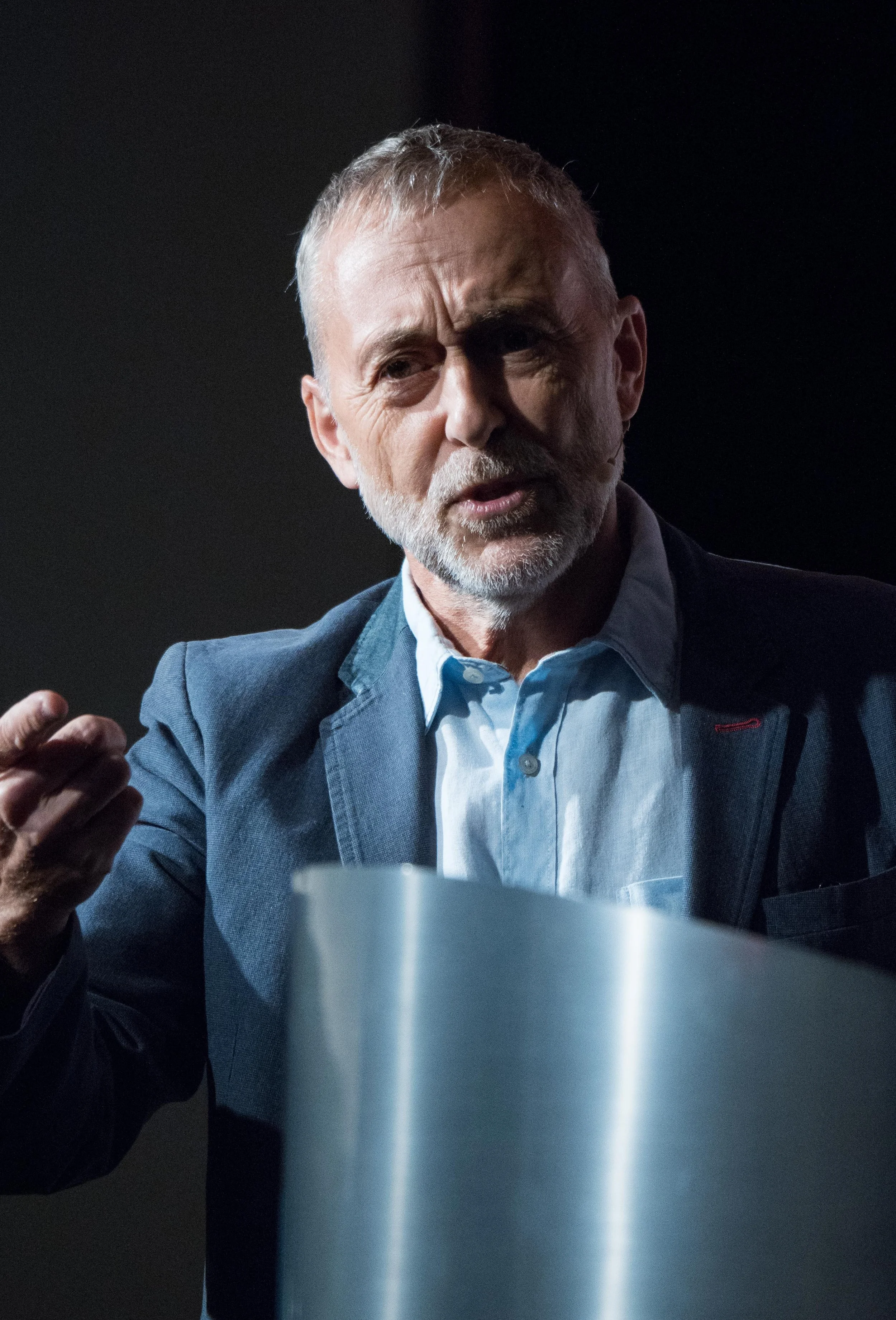 A man in a suit and dress shirt speaking at an event, with a dark background and a white cup in the foreground.