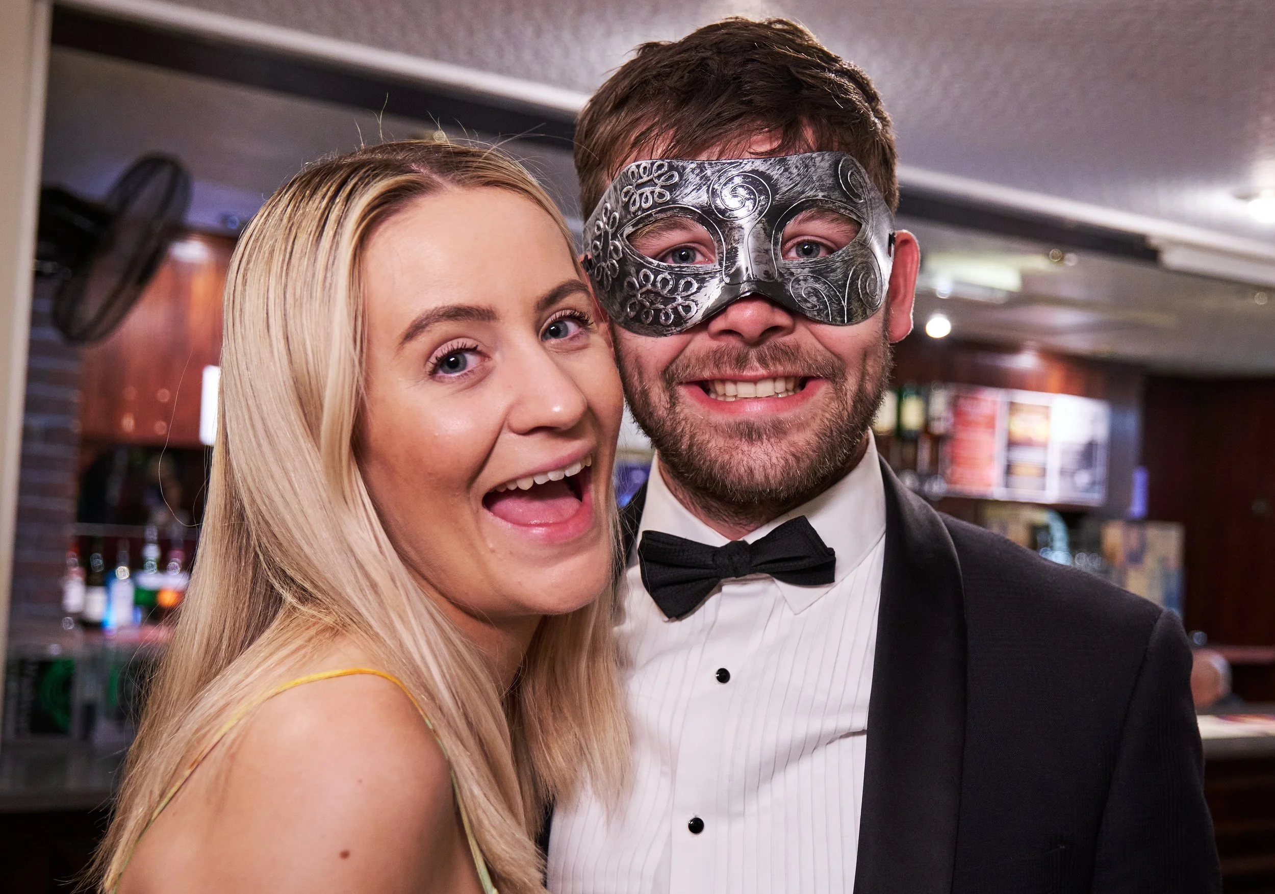 A smiling woman with blonde hair and a man with a beard, wearing a decorative metallic mask, dressed in formal attire at an indoor event.