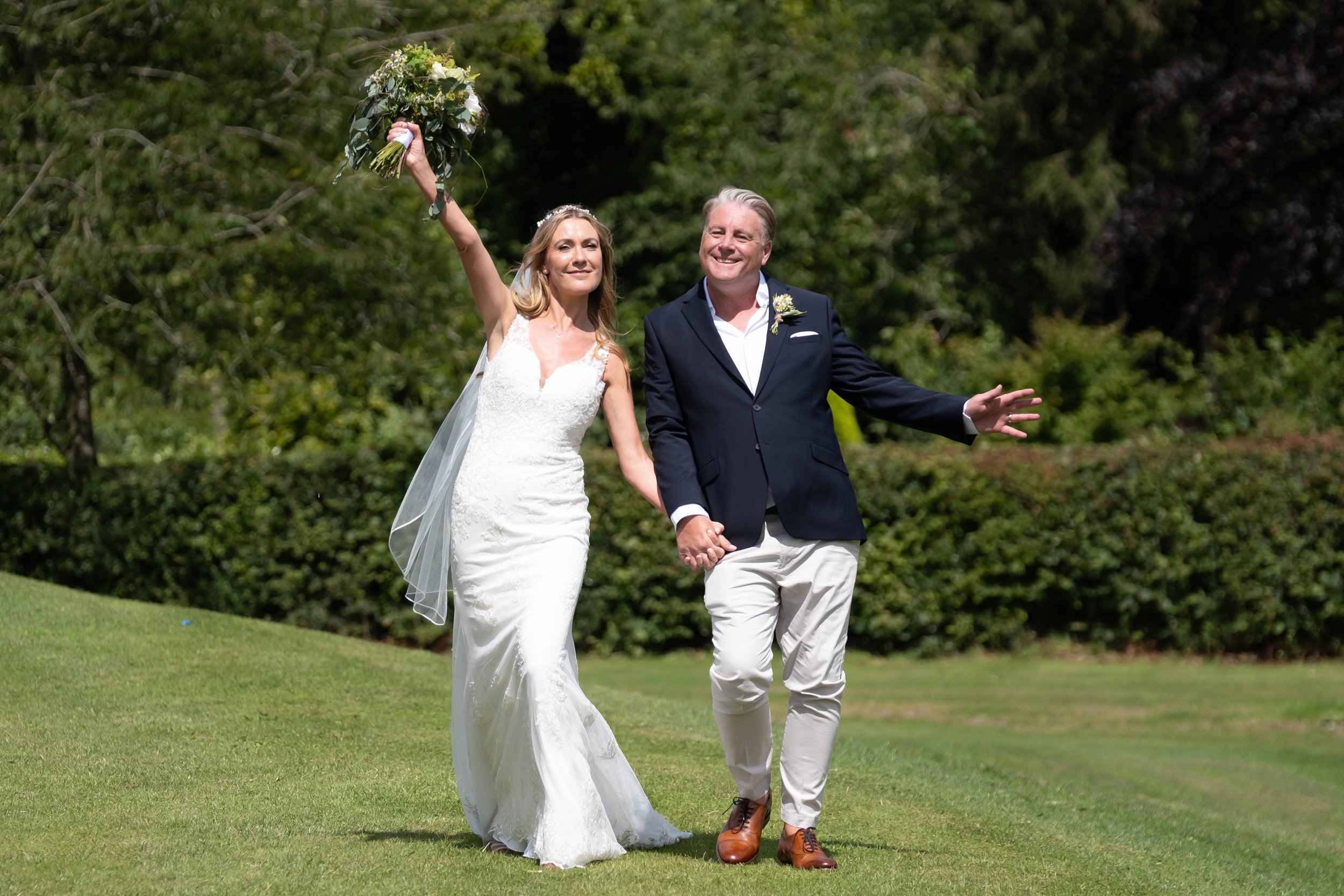 A bride and groom walking hand in hand outdoors on their wedding day, with the bride holding a bouquet of flowers in her raised hand and smiling.