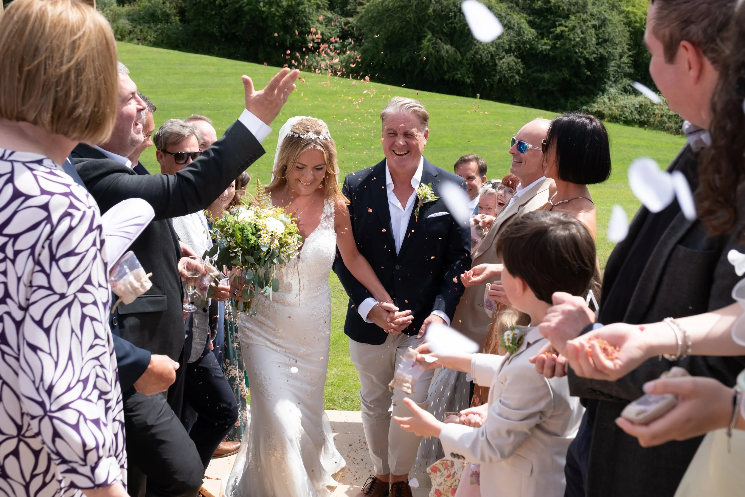 A bride and groom are smiling and holding hands outdoors at a wedding reception, surrounded by friends and family throwing flower petals and confetti on a sunny day with green grass and trees in the background.