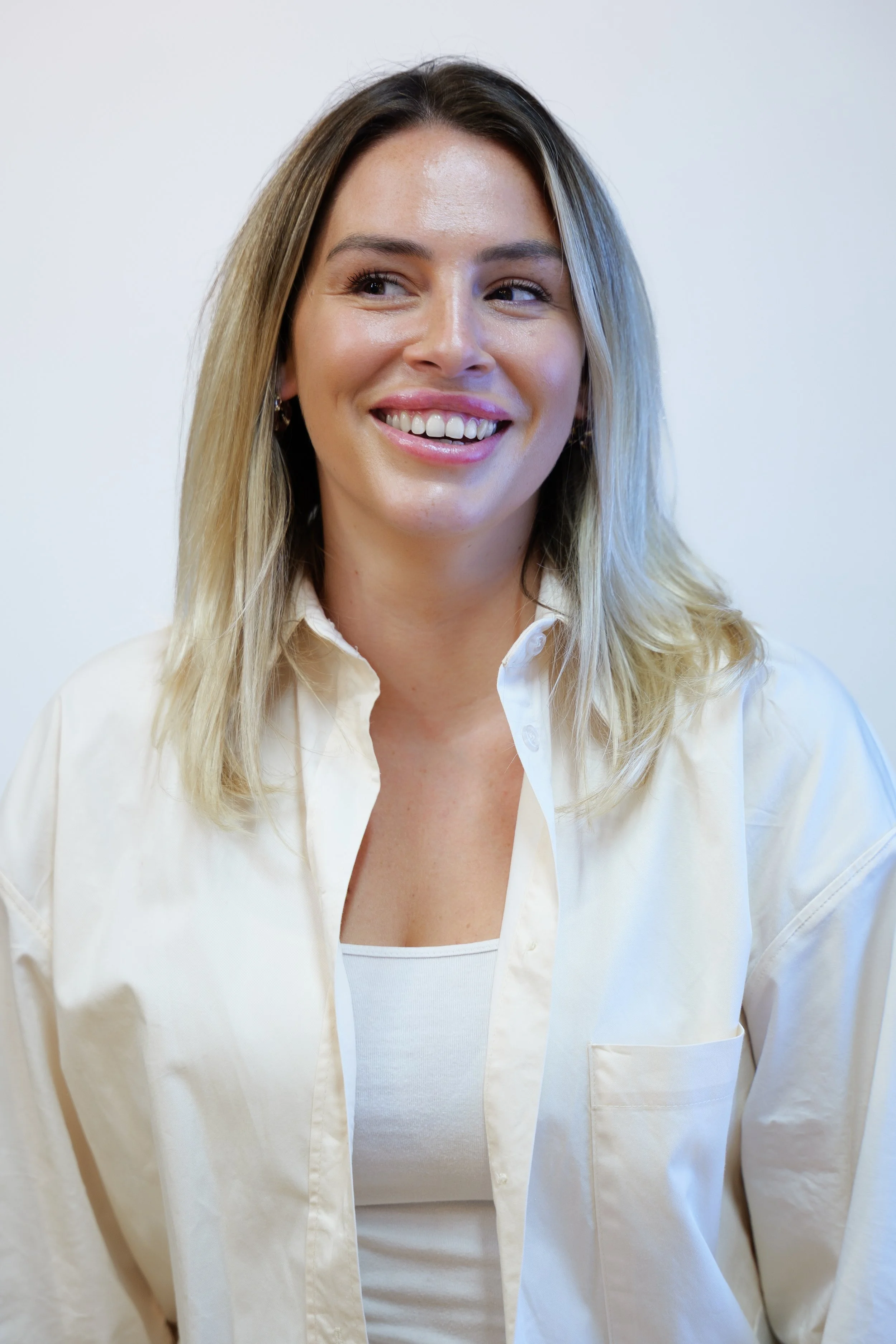 A smiling woman with long blonde hair, wearing a white shirt and tank top, standing against a plain white wall.