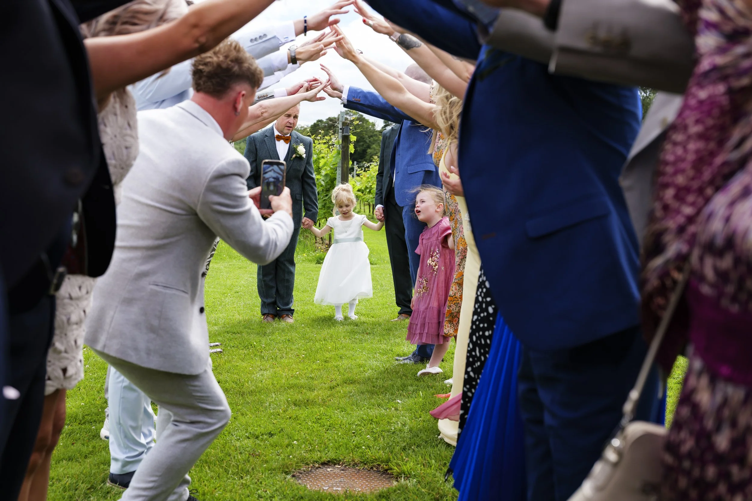 A bride and groom holding hands and walking together outdoors while surrounded by guests forming a pathway for them.
