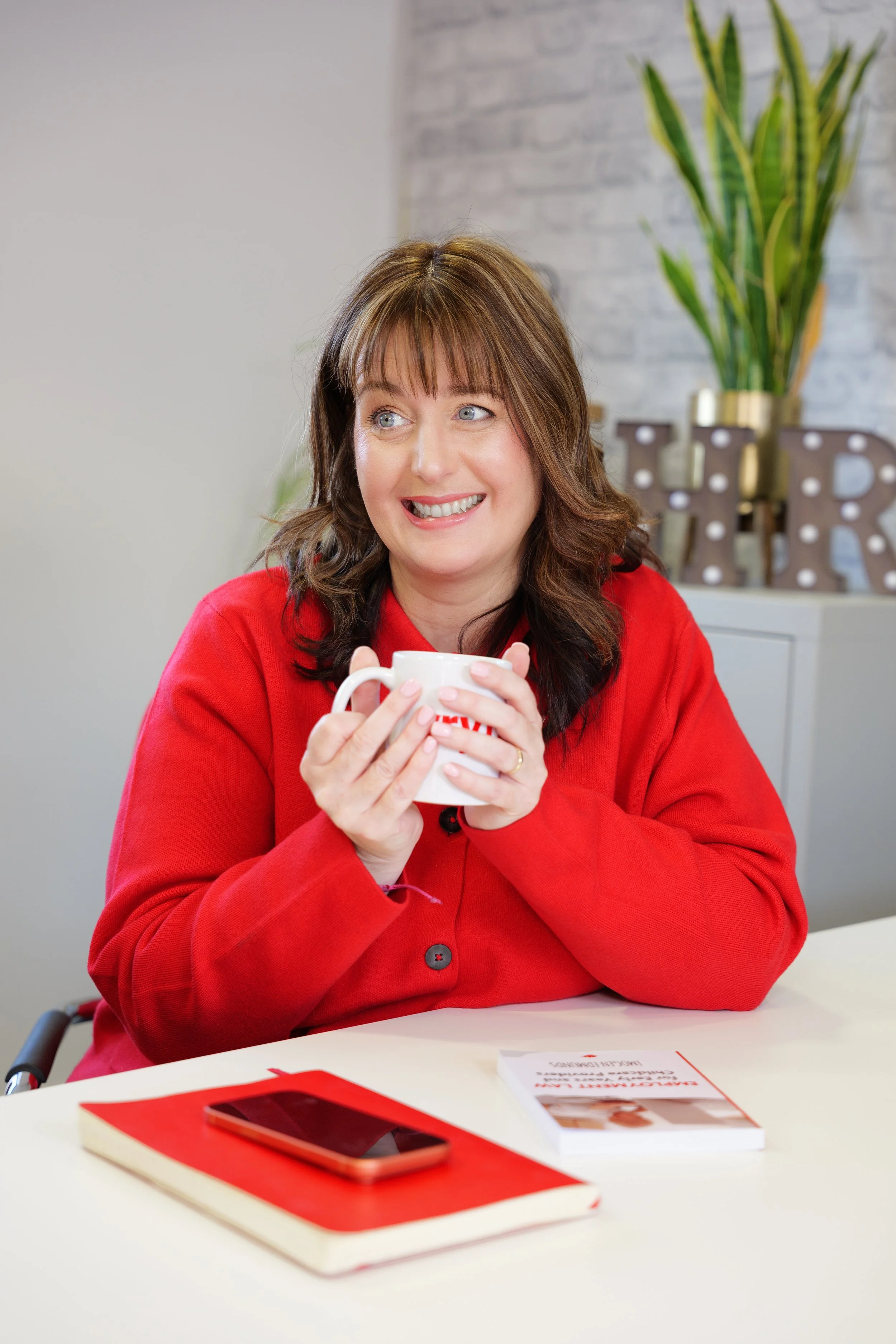 A woman with brown hair and blue eyes wearing a red blazer is sitting at a white table, smiling and holding a white coffee mug. On the table are a red notebook, a smartphone with a red case, and a brochure. There is a potted plant and decorative ligh