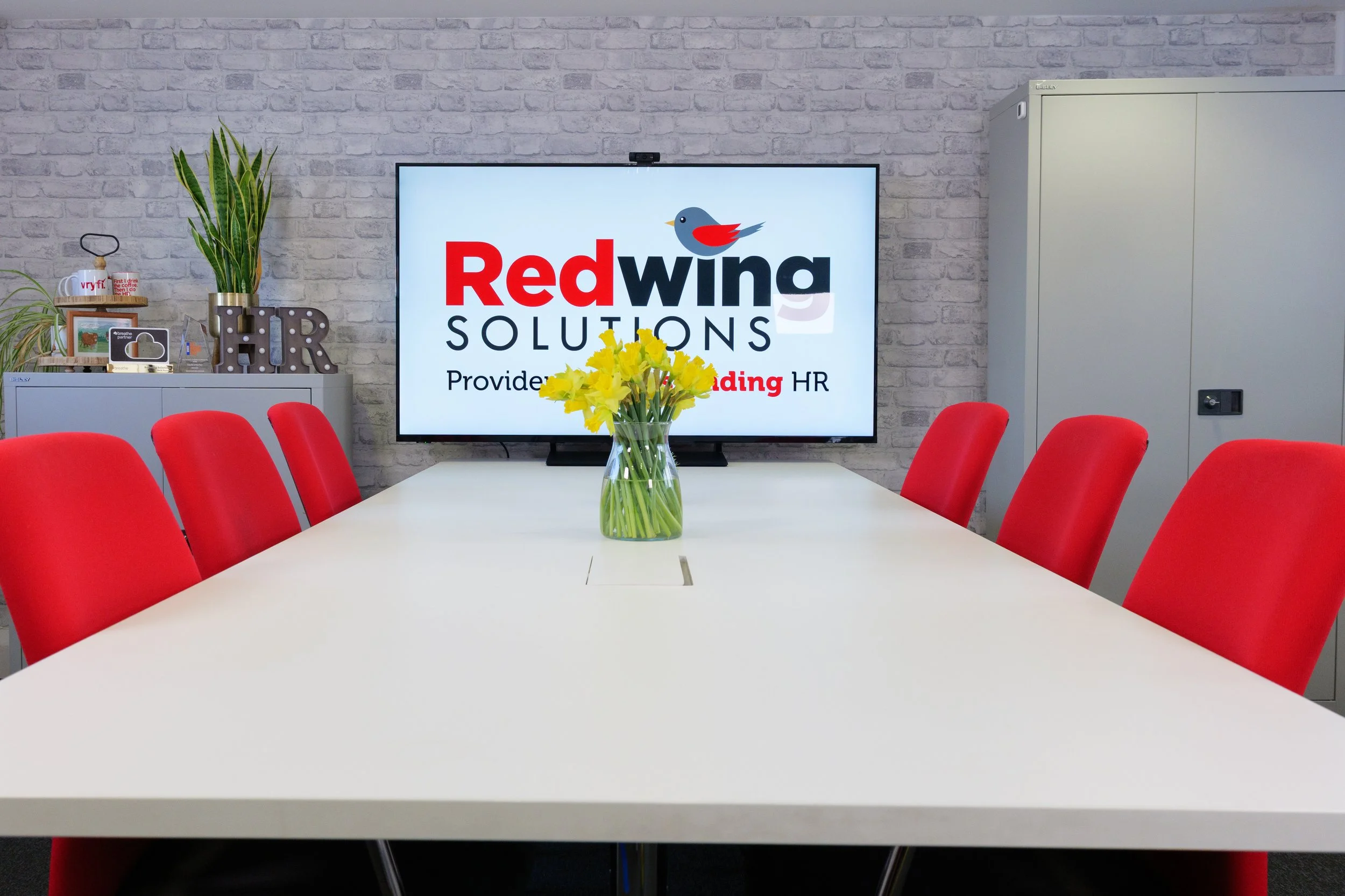 Conference room with a white table, red chairs, a vase of yellow flowers on the table, a large monitor on the wall displaying the Red Wing Solutions logo, and a gray cabinet with plants and decorative items.
