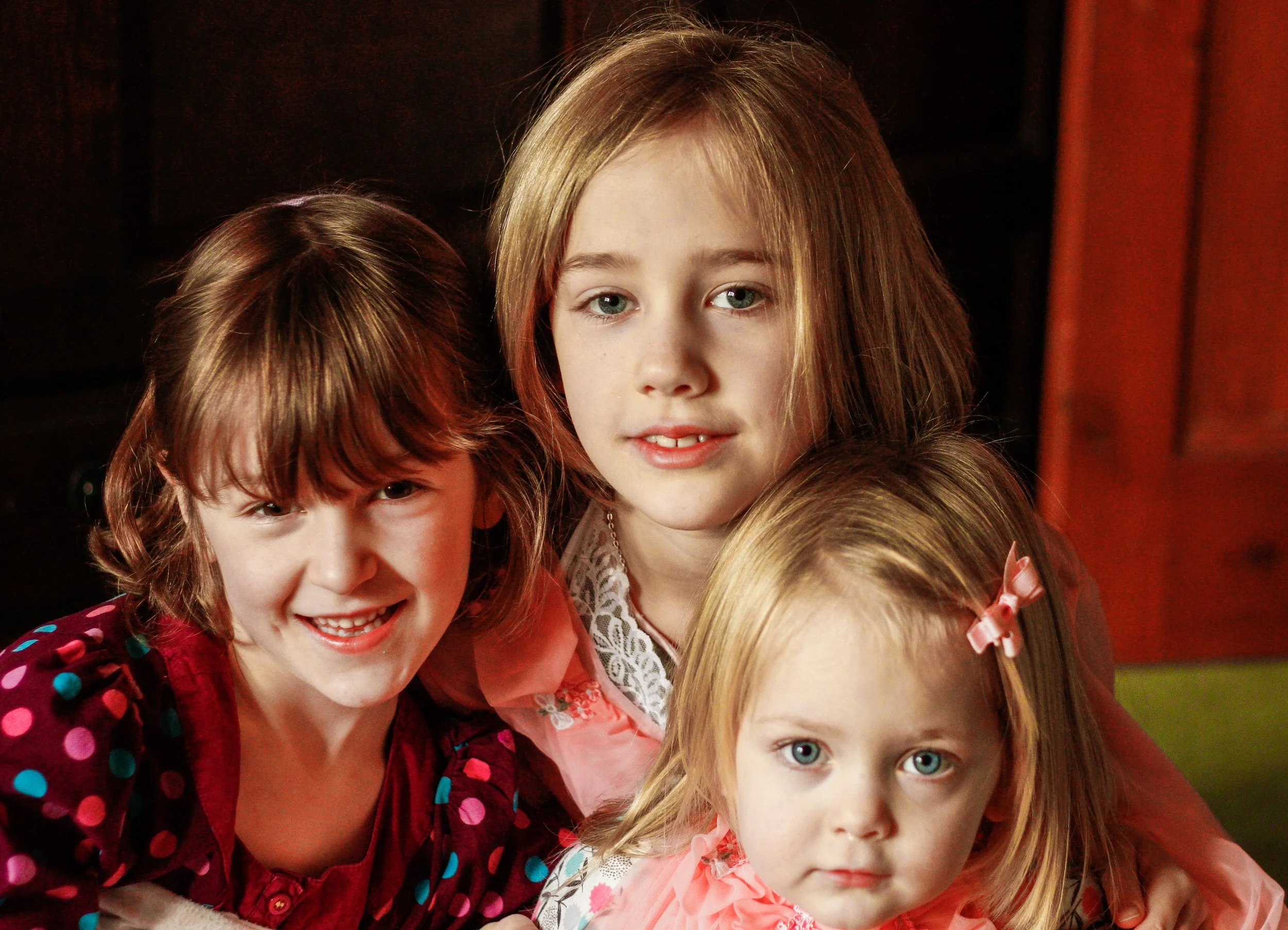Group of young girls with light hair and blue or green eyes, smiling and looking at the camera, dressed in colorful clothing, standing together indoors against a dark background with red curtains.