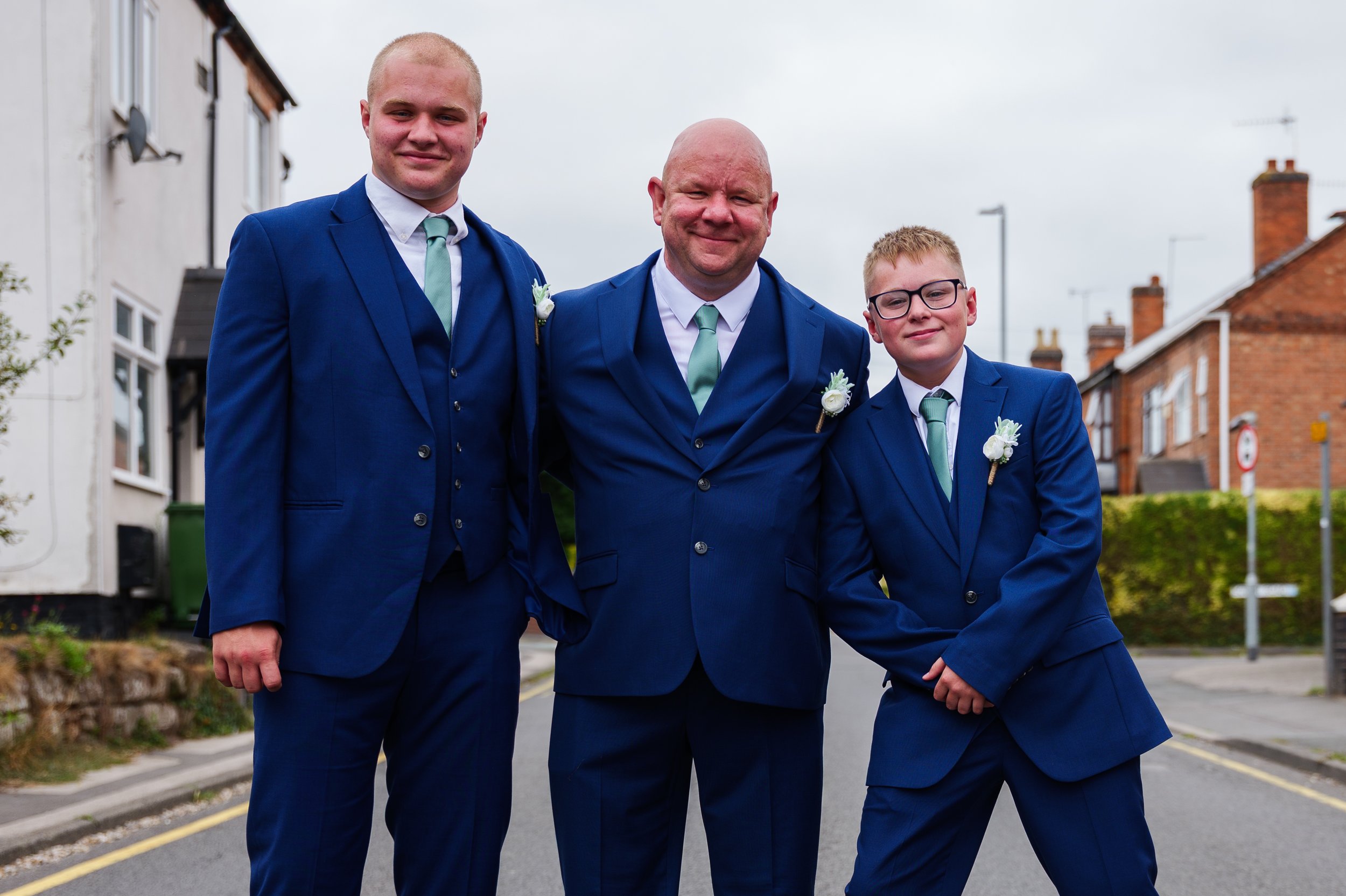 Three males in matching blue suits, white shirts, and mint green ties standing on a street with residential houses, all smiling.