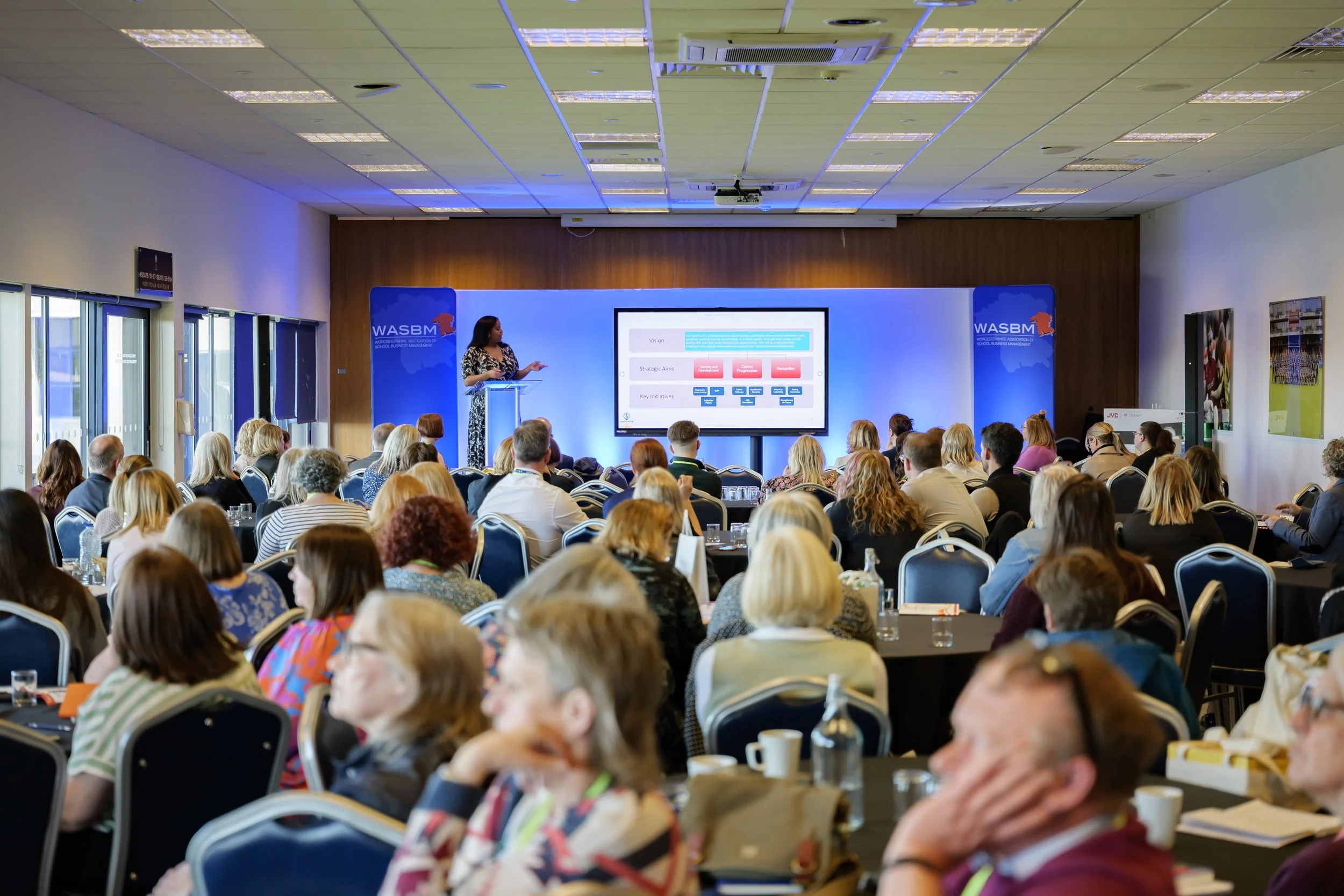 A woman giving a presentation in a conference room filled with an audience. The screen behind her displays a flowchart or diagram, and there are banners for WASBM on either side.