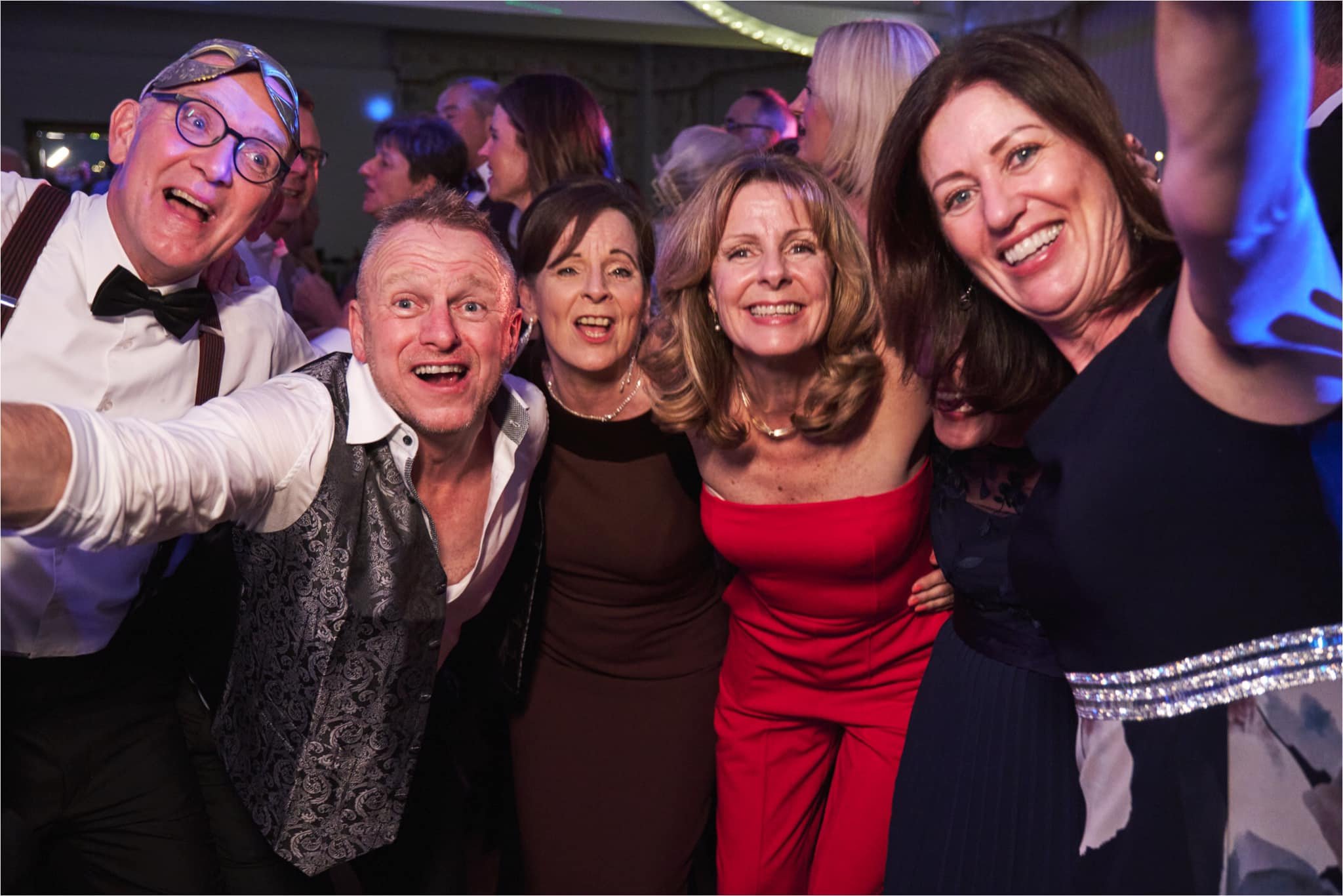 Group of people smiling and taking a selfie at a party or celebration, dressed stylishly with some wearing formal or semi-formal attire.