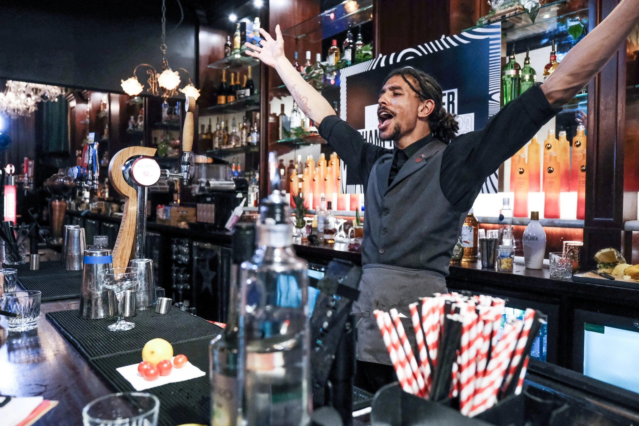 A bartender with dreadlocks and a black shirt is raising both arms and singing or speaking passionately behind a bar, surrounded by liquor bottles and bar tools.