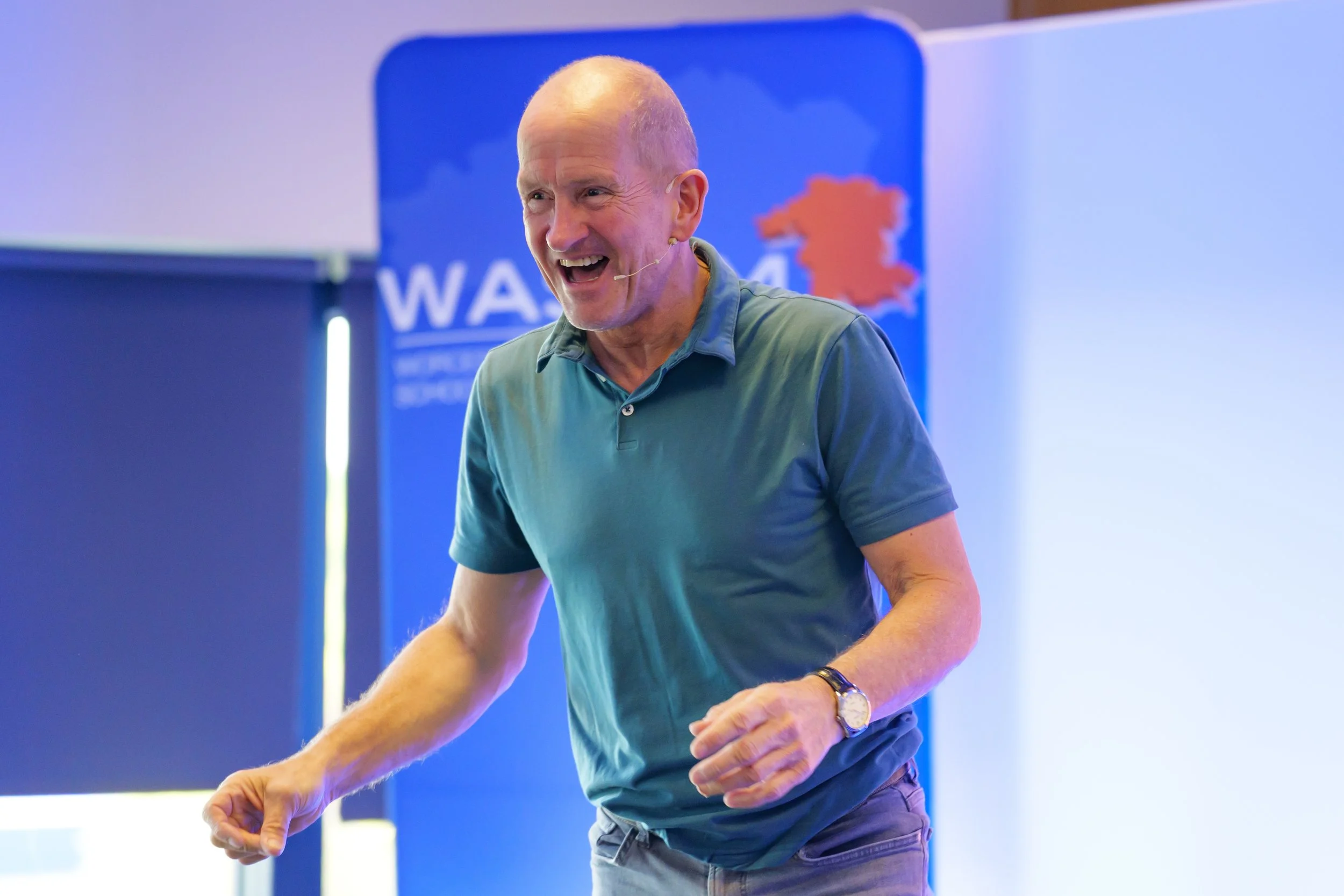 A middle-aged man with a shaved head smiling and speaking into a headset microphone during a presentation, standing in front of a blue background with a logo.