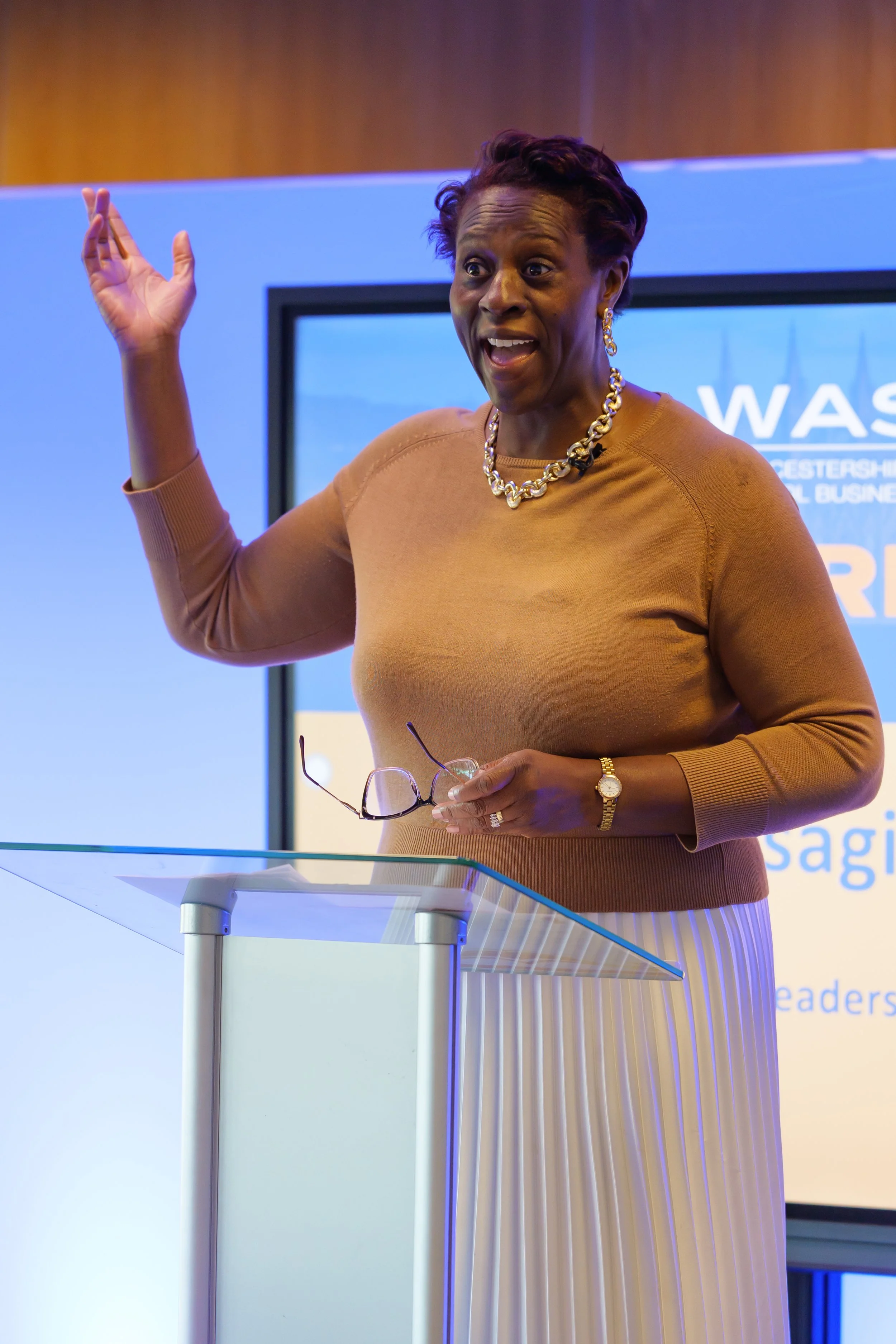A woman speaking at a podium with a modern background, wearing a tan sweater, striped skirt, gold necklace, and holding glasses.